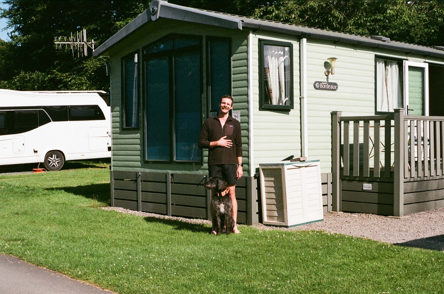 A man in a dark fleece and shorts stands beside a black curly-haired dog on the grass in front of a green static caravan. A white campervan is parked to the left.