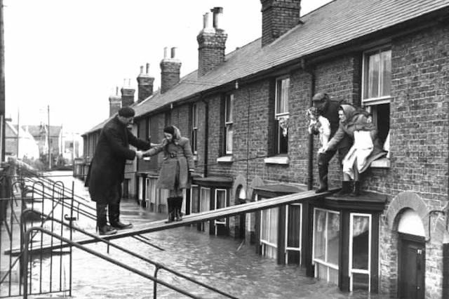 A historic black-and-white photo showing people navigating a flooded street; one person extends a hand to help a woman across a makeshift bridge, while others watch from windows.
