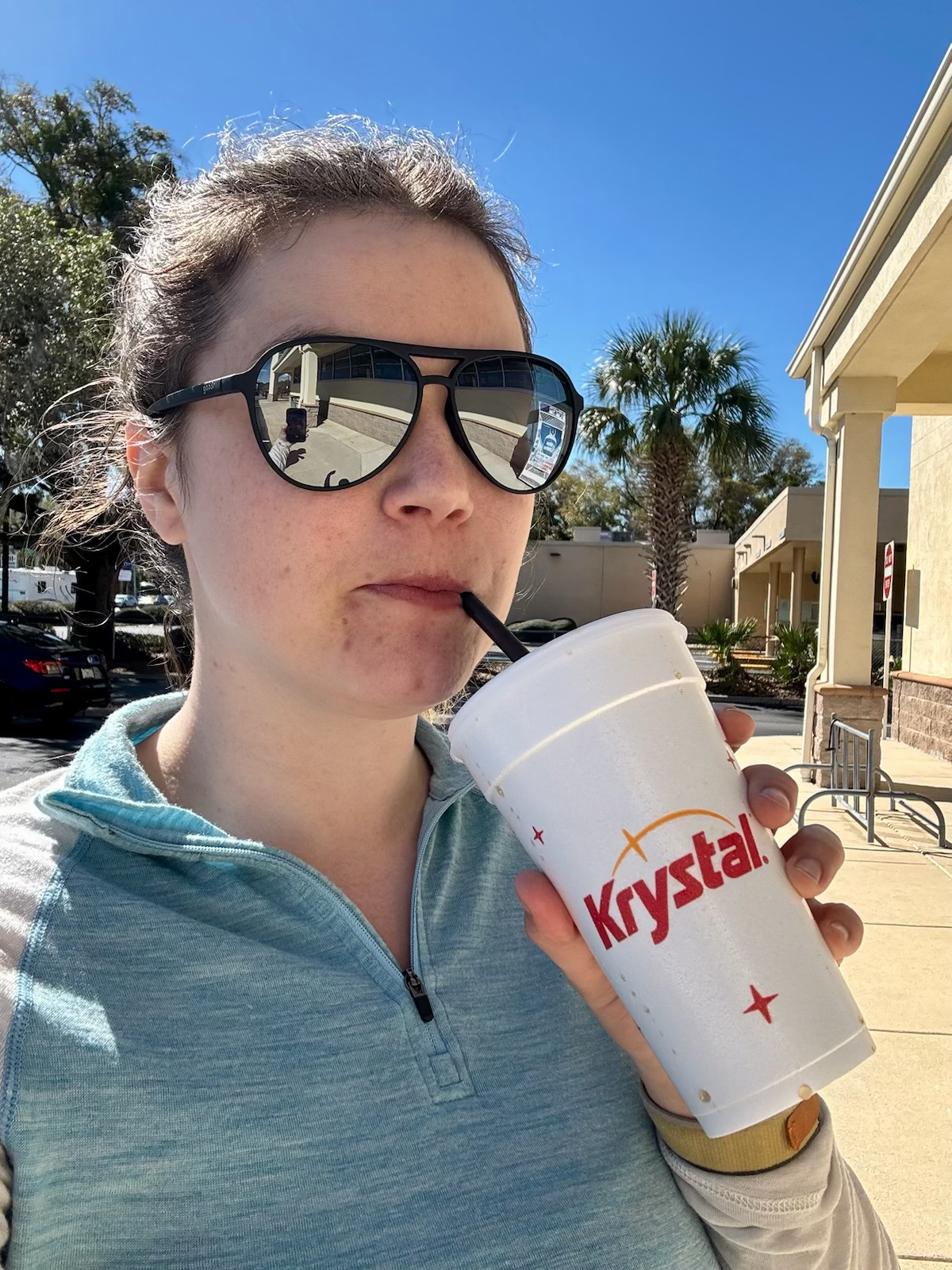 A woman in aviators drinking out of a polystyrene cup in front of some palm trees.