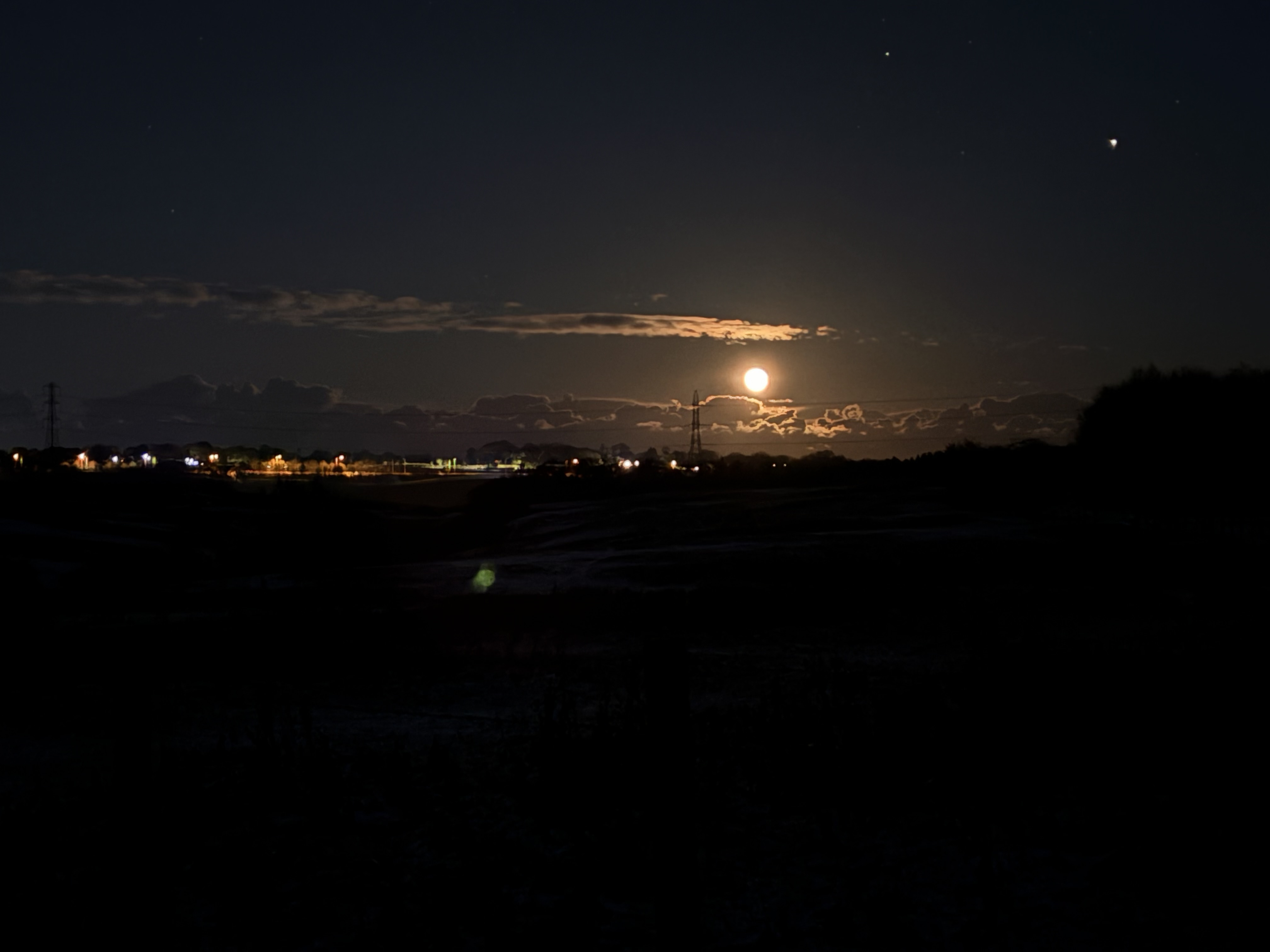 A bright moon above clouds on the horizon, some far off lights in the distance, all dark close to the camera