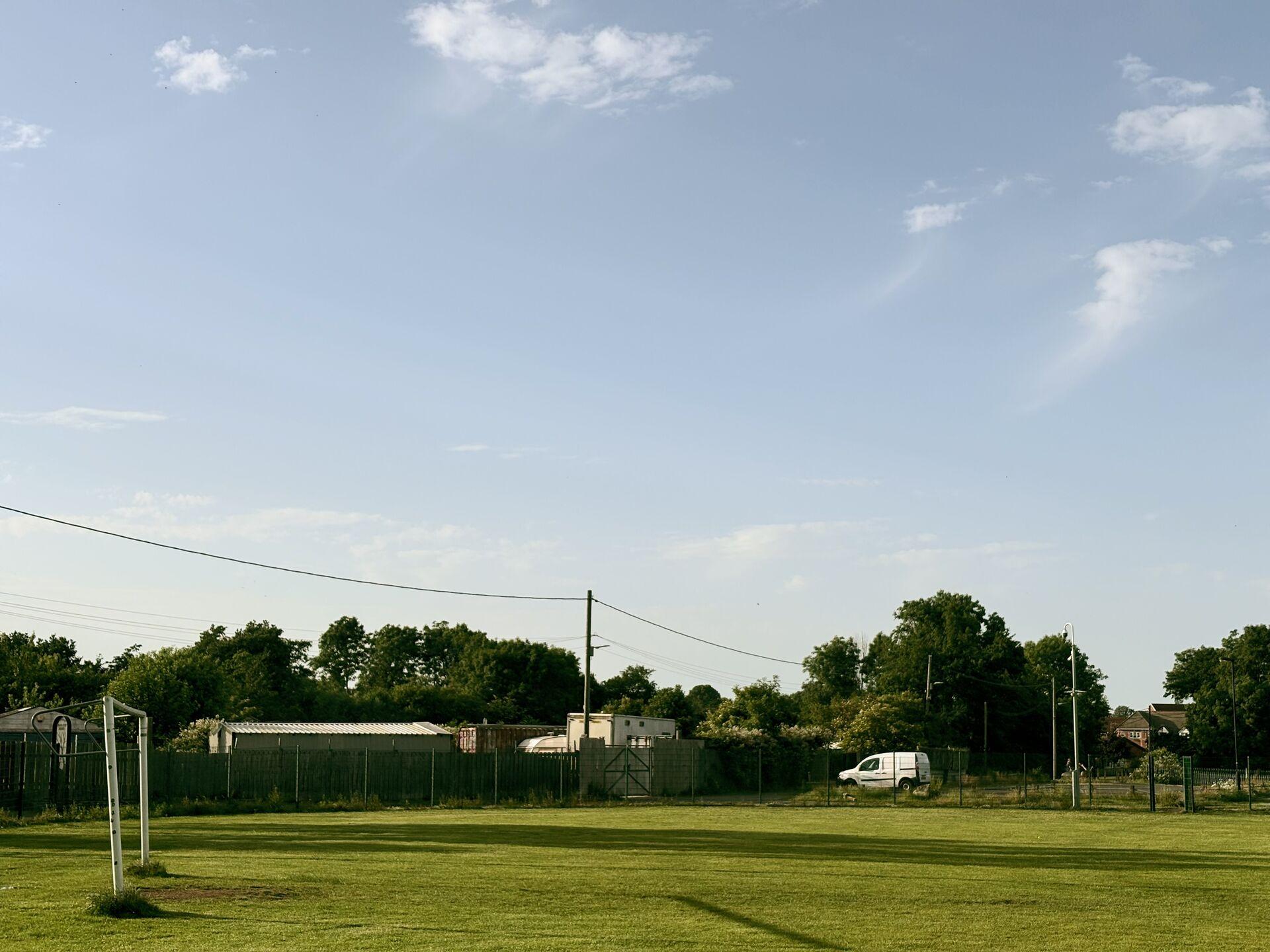 A mowed grass pitch with a goalpost visible on the left, backed by a chain-link fence, outbuildings, and a row of trees under a blue sky with light cloud.