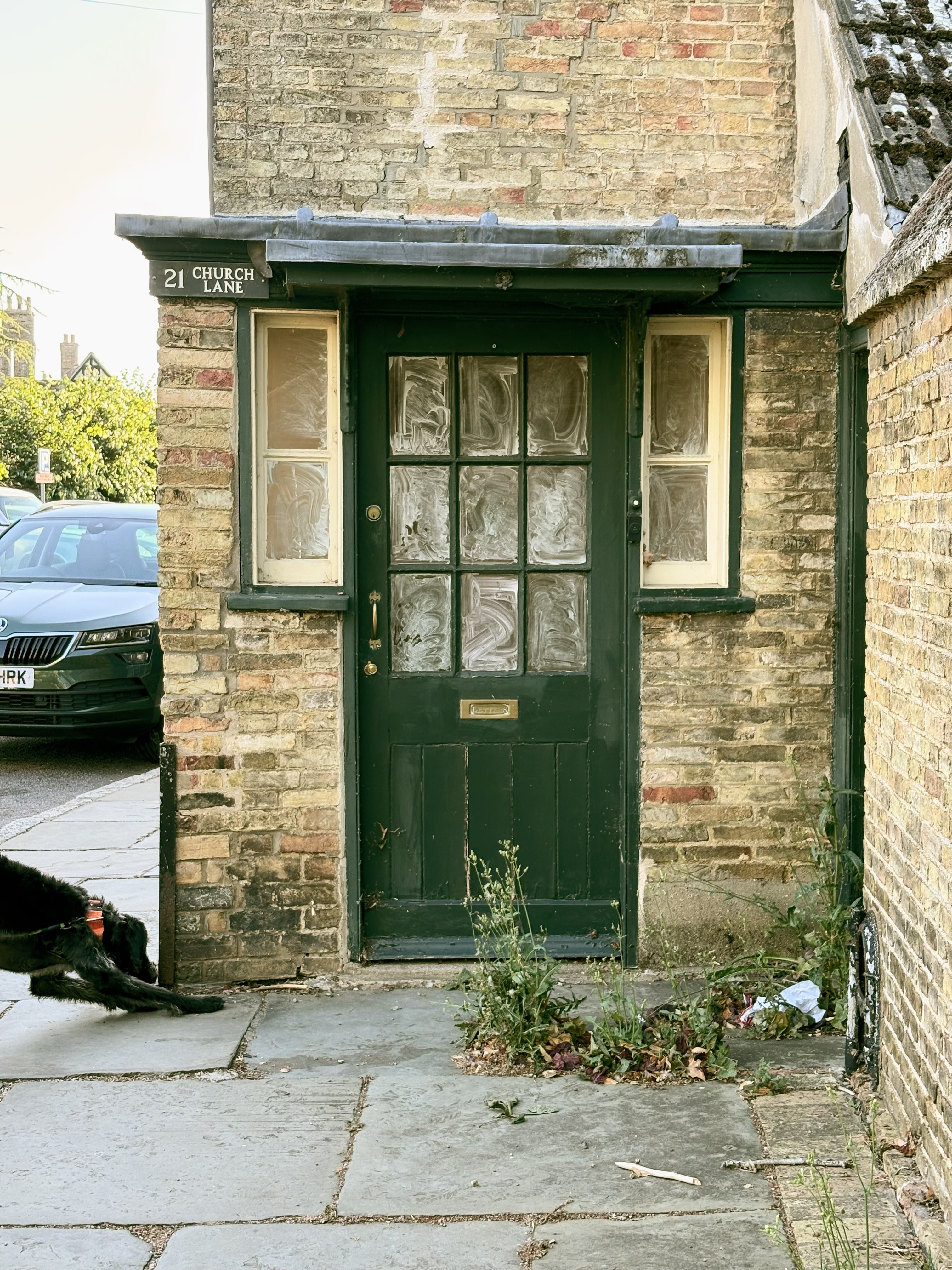 A square-on view of a black windowed door set in a yellow brick wall under a tile roof overgrowing with moss.