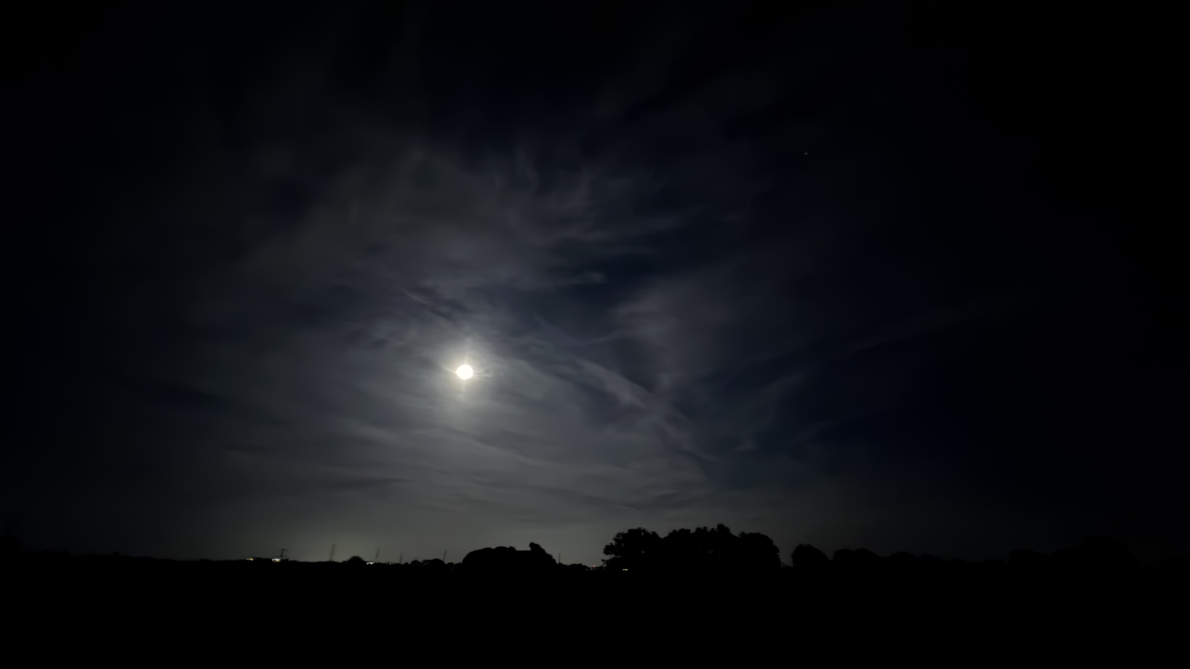 A wide angle of a dark sky with a bright moon in the middle of it. The outlines of trees along the horizon are visible at the bottom of the frame.