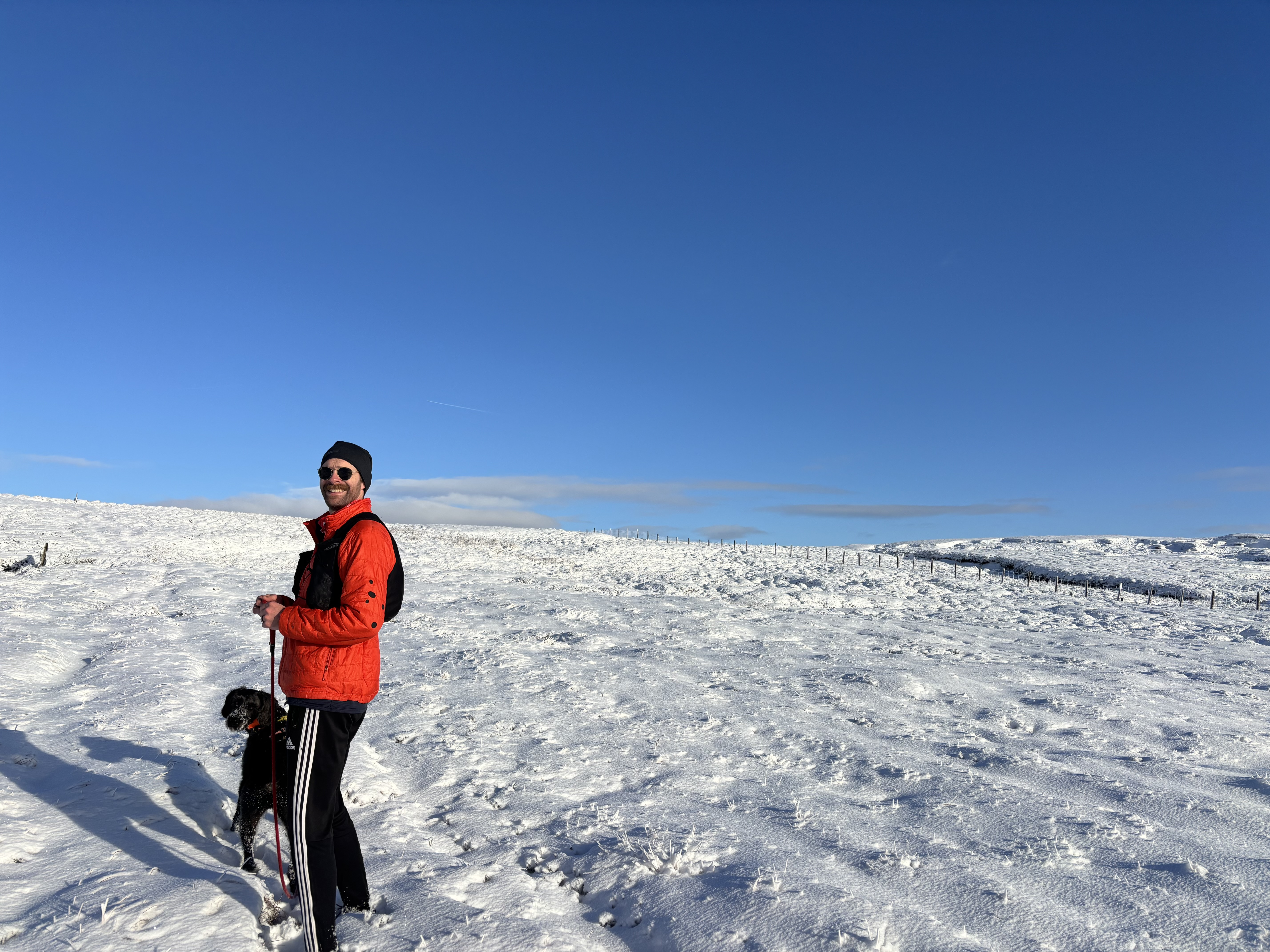 A man in a bright orange jacket, dark tracksuit pants, beanie, and sunglasses stands on a snowy hillside, with a dog at his side