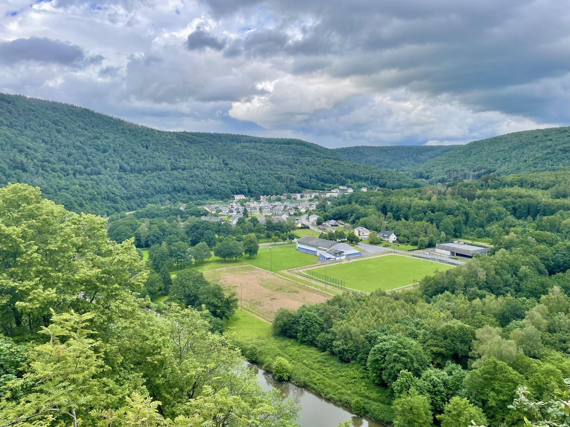 The view from above the town of Les Hautes Rivières, looking down on rolling hills covered with trees and some grey buildings arranged at the bottom of the valley, under a looming sky