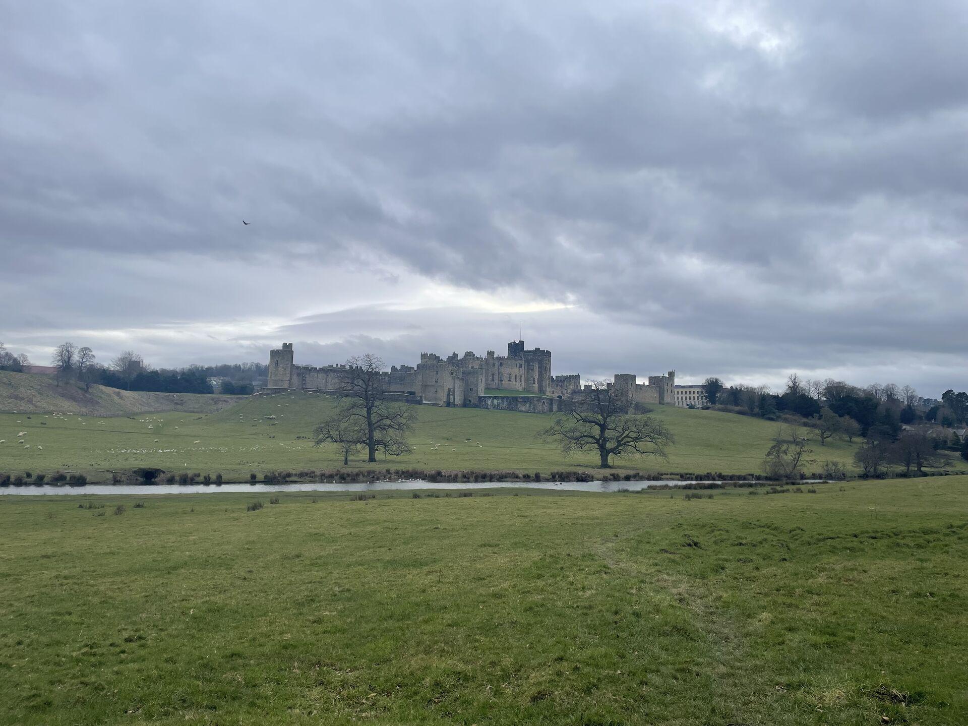 Historic Alnwick Castle sits atop a grassy hill, surrounded by trees and a river in the foreground under a cloudy sky.