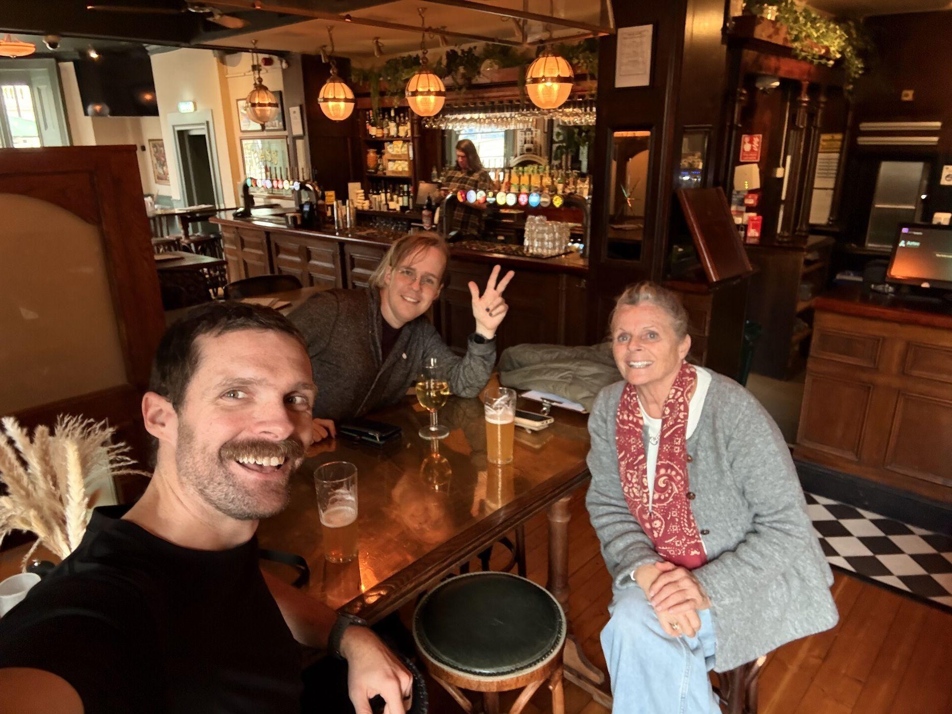 A selfie of two men and a woman in a pub, with drinks on the table and a checkerboard floor in front of the bar in the background