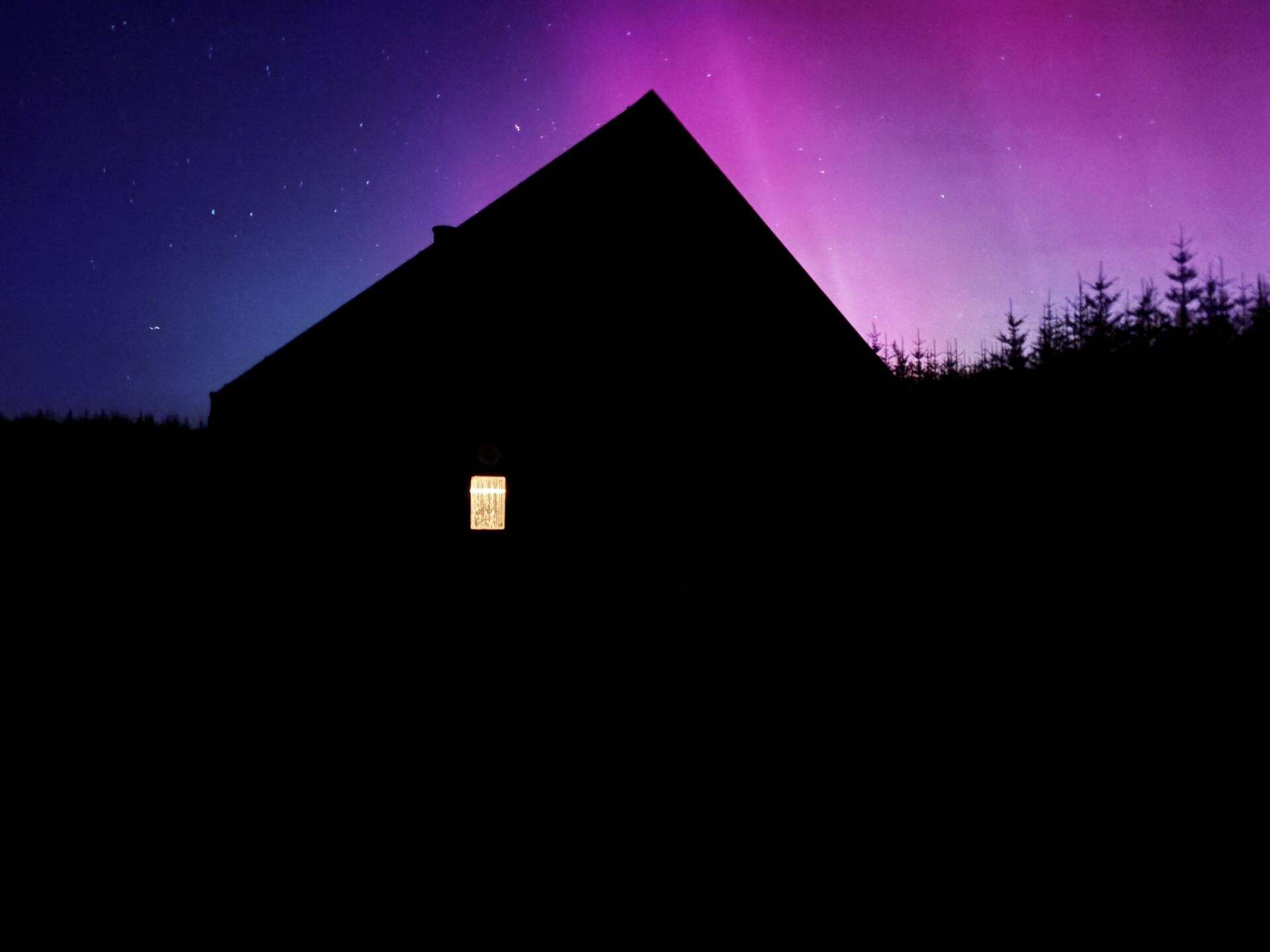 A purple aurora above the silhouette of a bothy in the woods