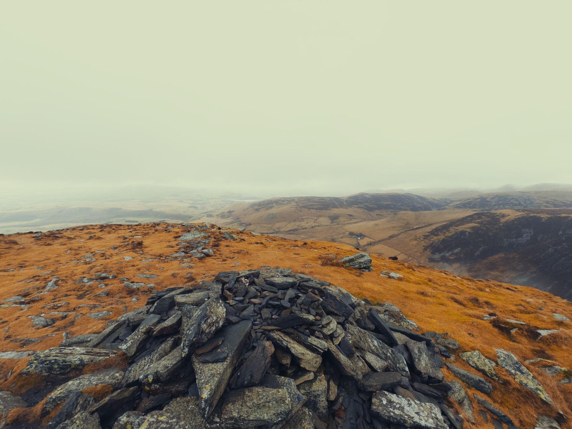 A low shale cairn on a grassy summit, low heather-covered hills stretching off into the hazy distance.