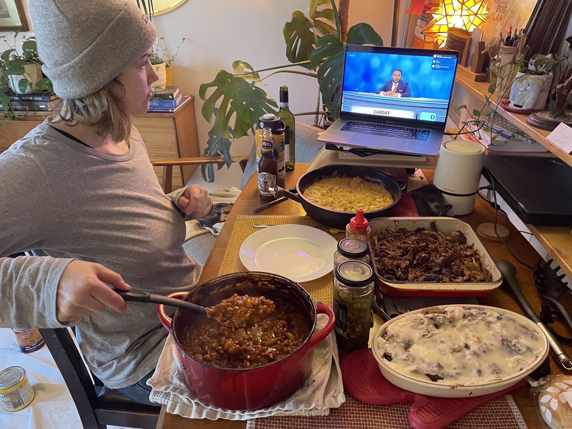 A table set with mac n cheese, pulled pork, bbq beans, and bread pudding, with a woman sat at the side of the table spooning out beans and watching University Challenge on a laptop