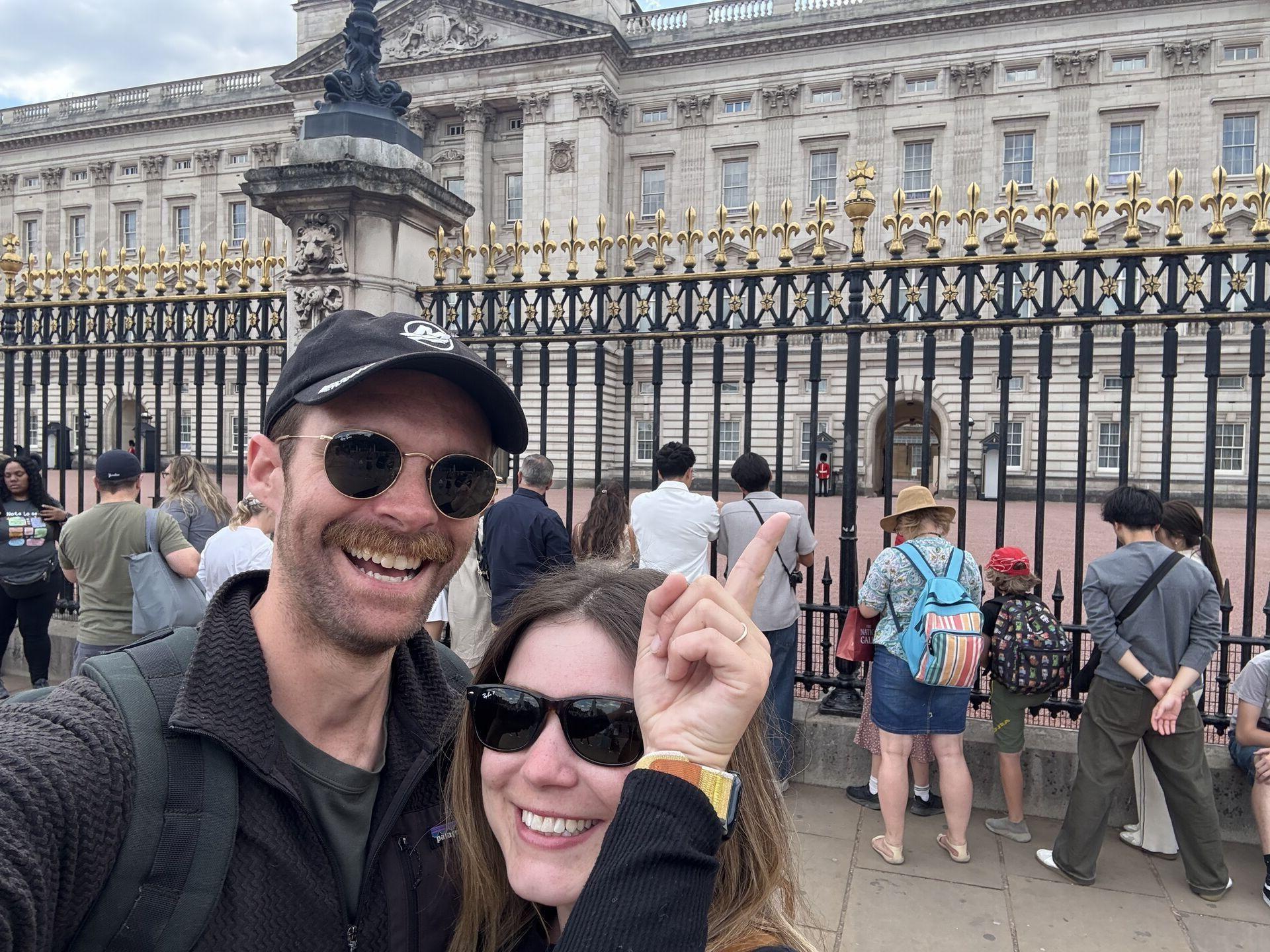 Two smiling tourists wearing sunglasses take a selfie in front of the gated Buckingham Palace, while a crowd stands behind them looking at the palace. One points towards a palace guard.