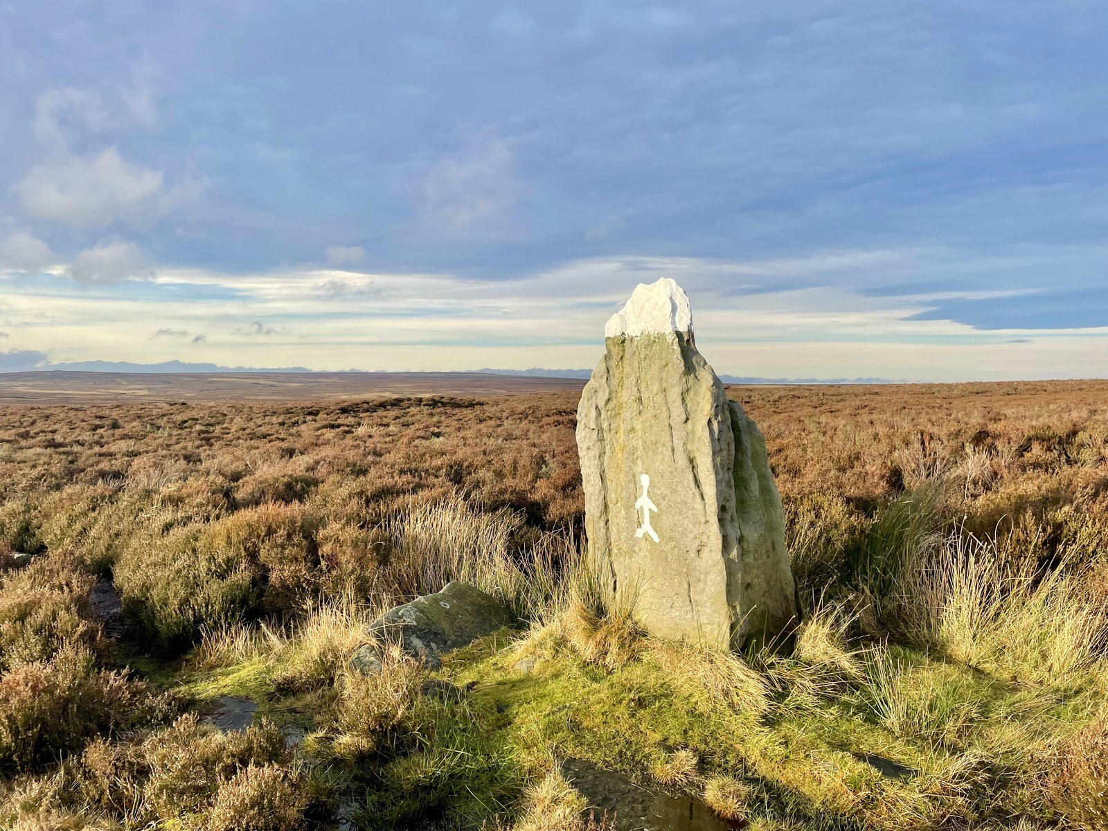 A massive standing stone, capped with white paint and with a stick figure graffito, on a massive empty heather moor