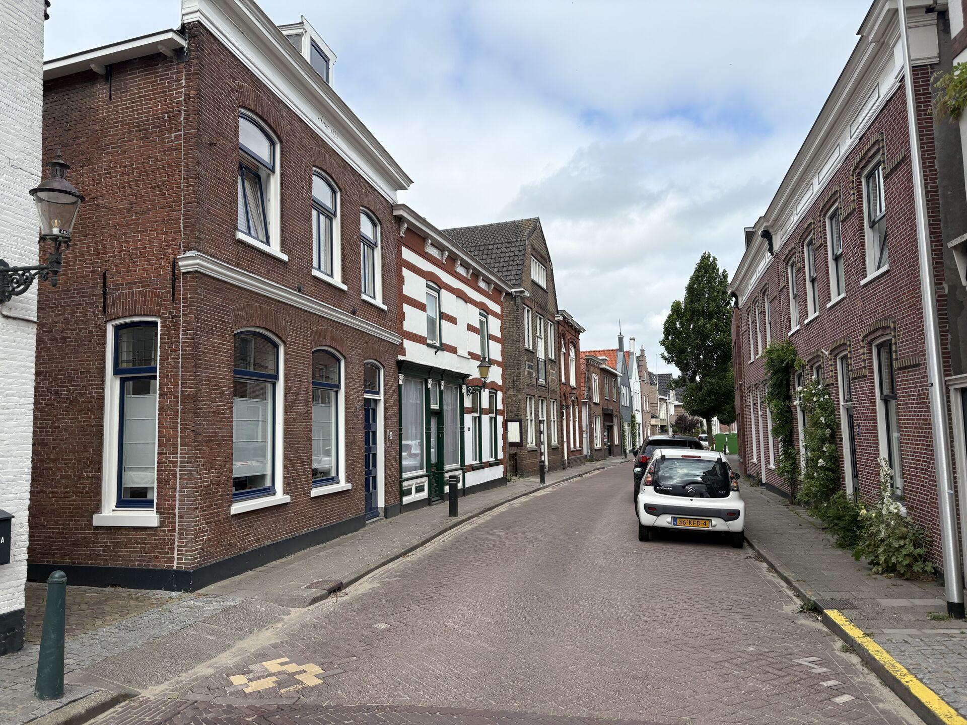 Street view of a narrow European road lined with brick buildings, cars parked on the right side, a lamppost on the left, and a cloudy sky overhead.