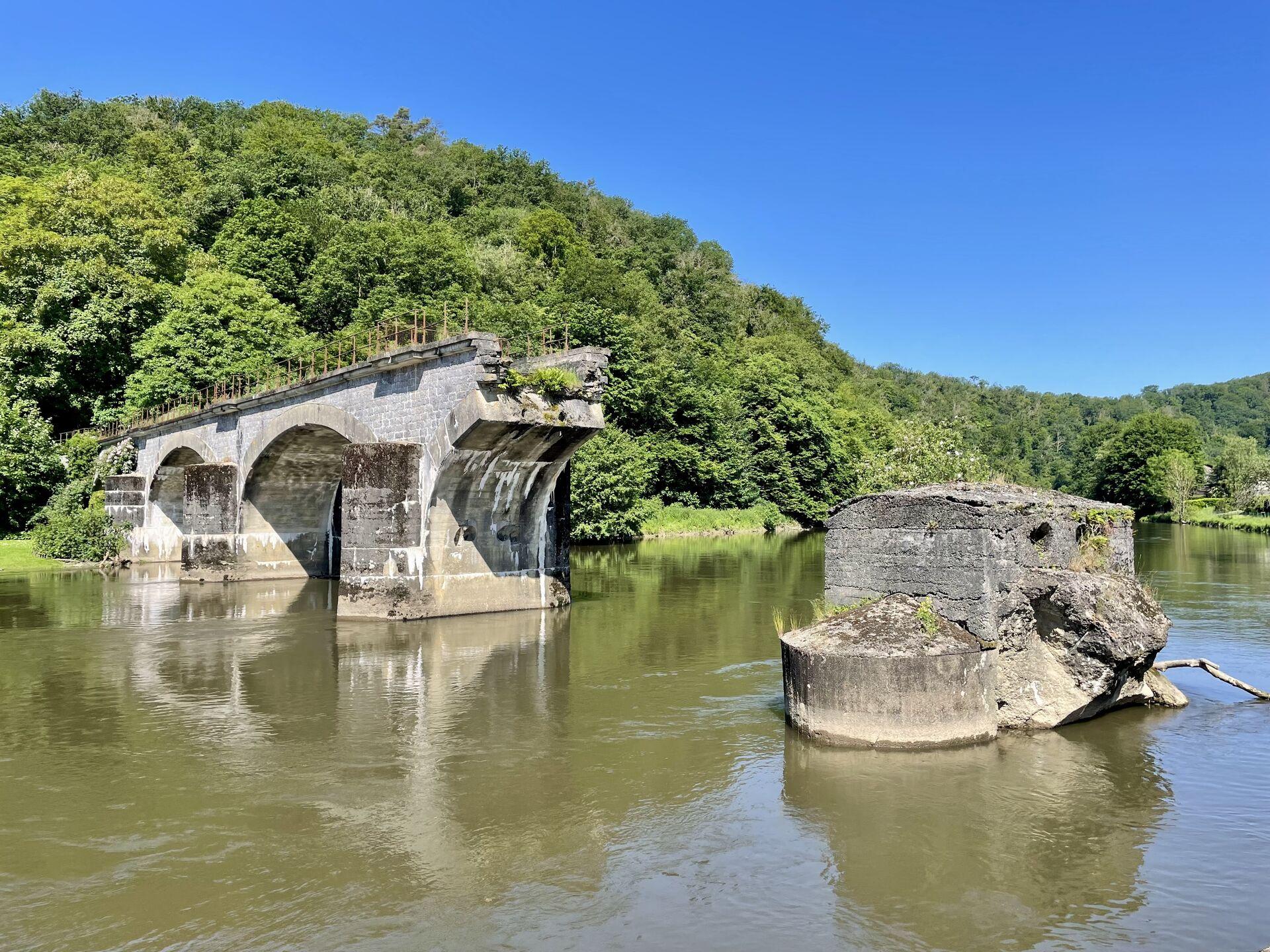 A demolished bridge, with two arches intact and two arches missing, over a greenish river with a sunny forested hillside rising behind