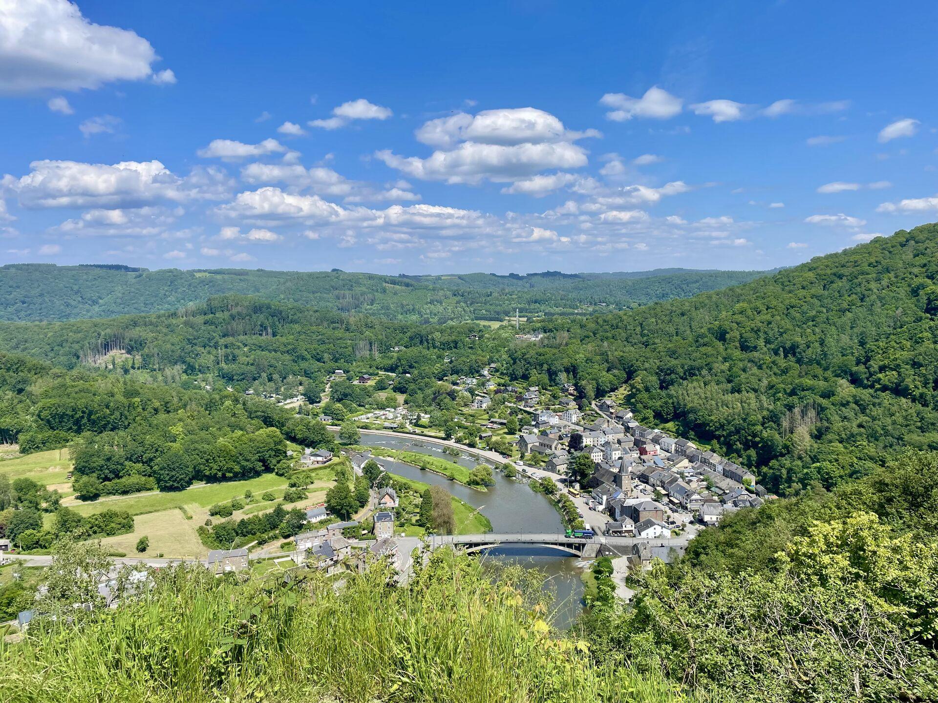 A view of a little grey village nestled in the bend of a river, surrounded on all sides by green hillside, under a blue sky with scattered clouds