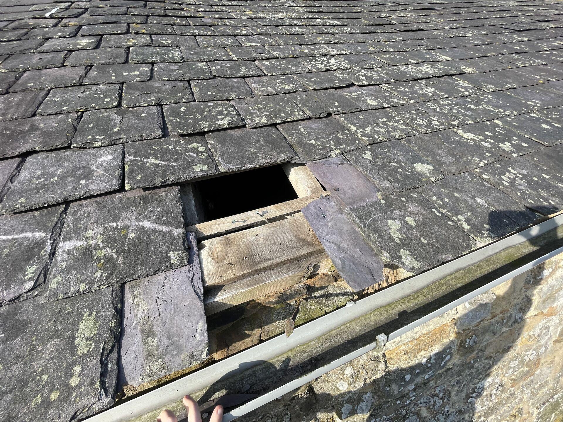 A close-up view of a slate roof with a visible hole, revealing wooden beams underneath.