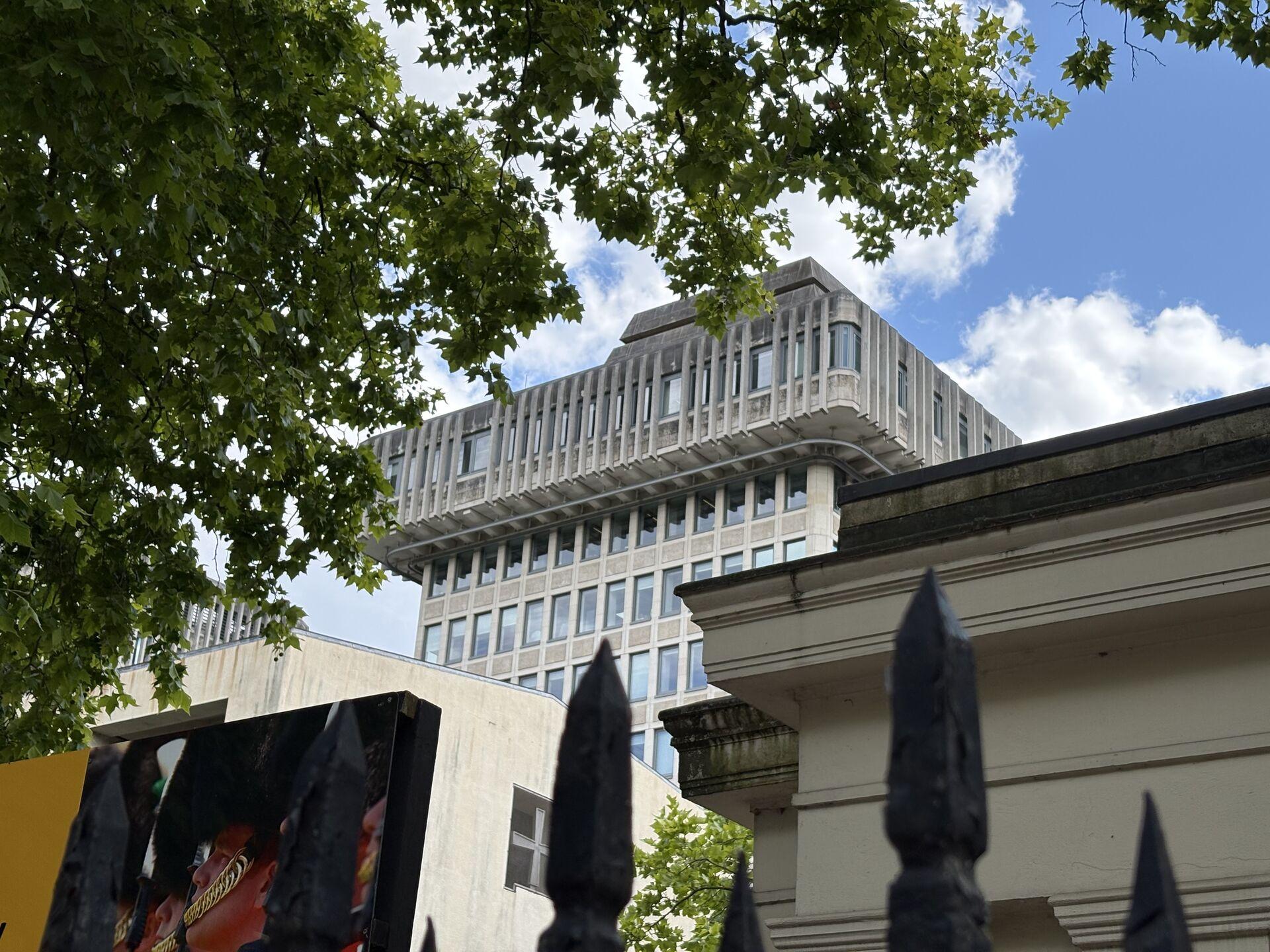 View of a tall, gray, brutalist-style building partially obscured by green leaves, a white building, black iron fence spikes, blue sky, and clouds.