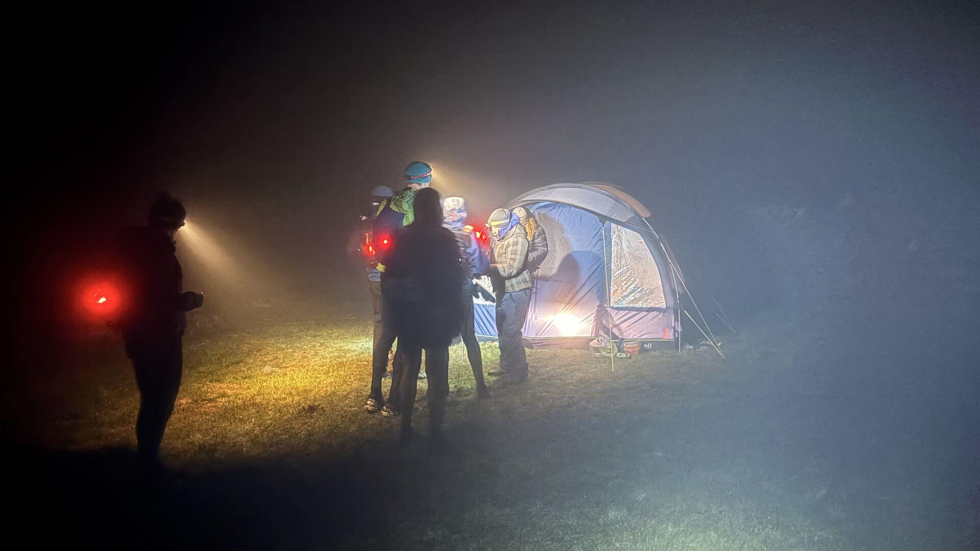 A handful of people standing by a tent in the dark, illuminated by each other's headtorches; one is getting their tally clipped by a marshal