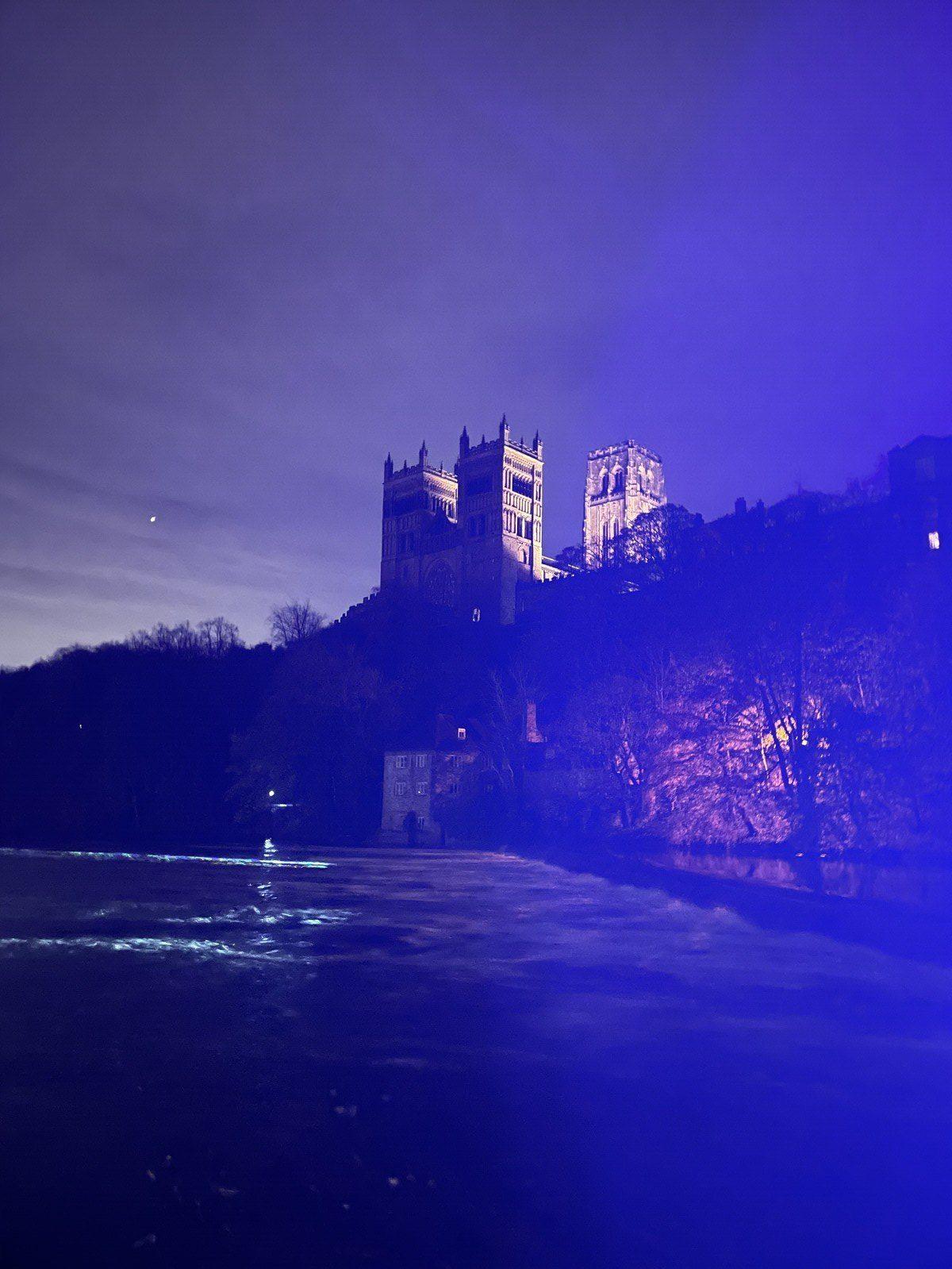 Durham Cathedral viewed from below, with the River Wear in the foreground, illuminated by a blue glow from somewhere off to the right
