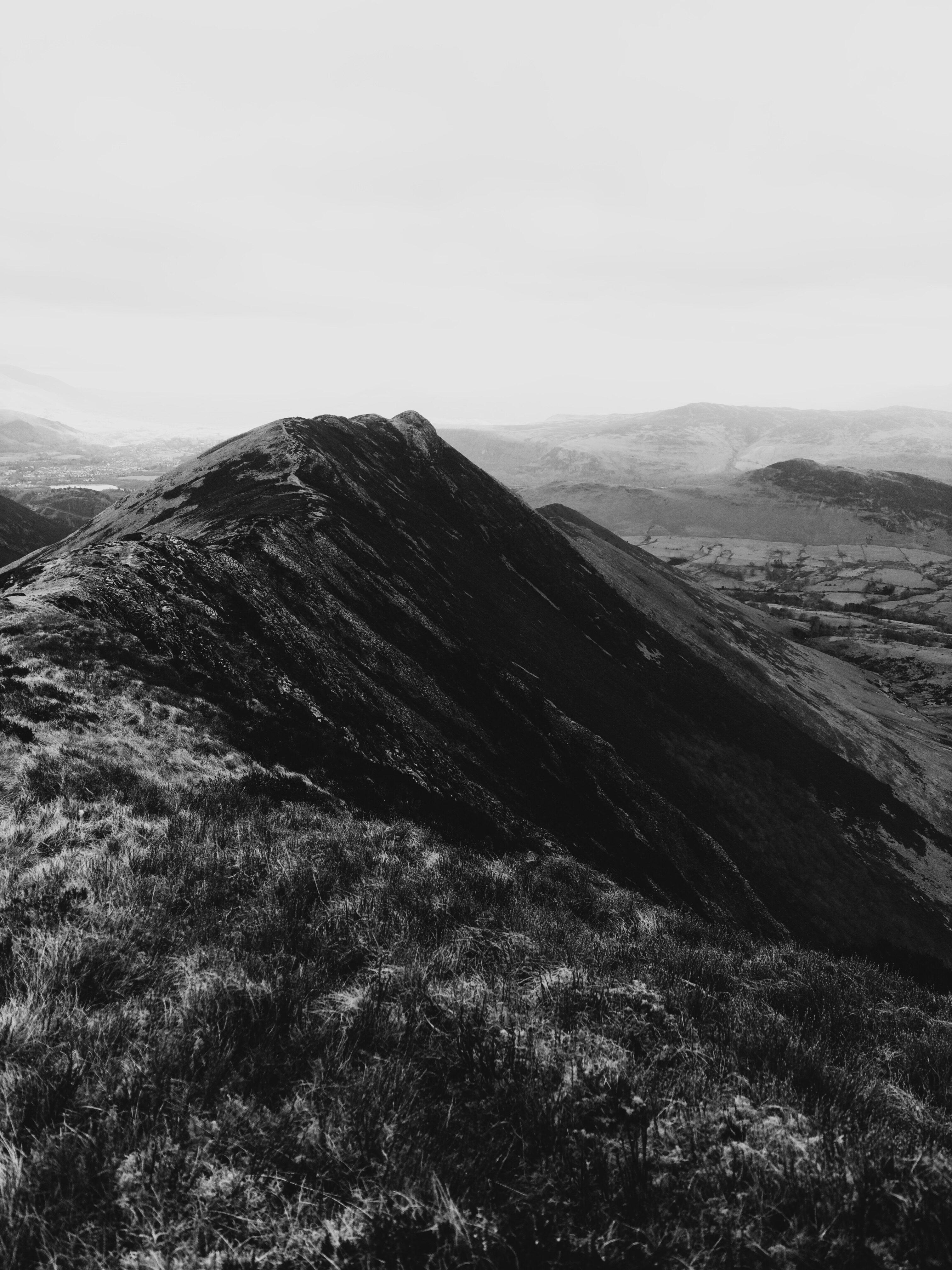 A dramatic black and white photo of the side of a fell in the lake district, with crags and scree running off the right side and some blown-out fells fading off in the background