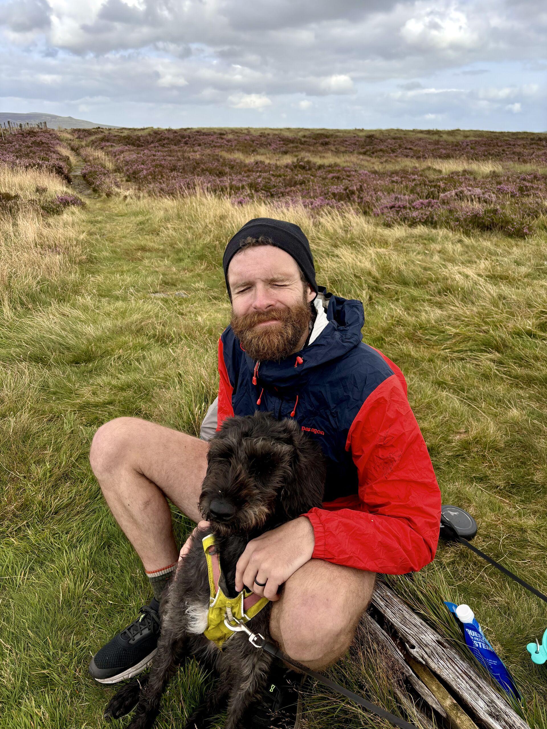 Me, sitting on a log on in a grassy field surrounded with heather, wearing a windbreaker and toque, squinting at the camera with a black dog sitting in between my legs, looking at something out of frame