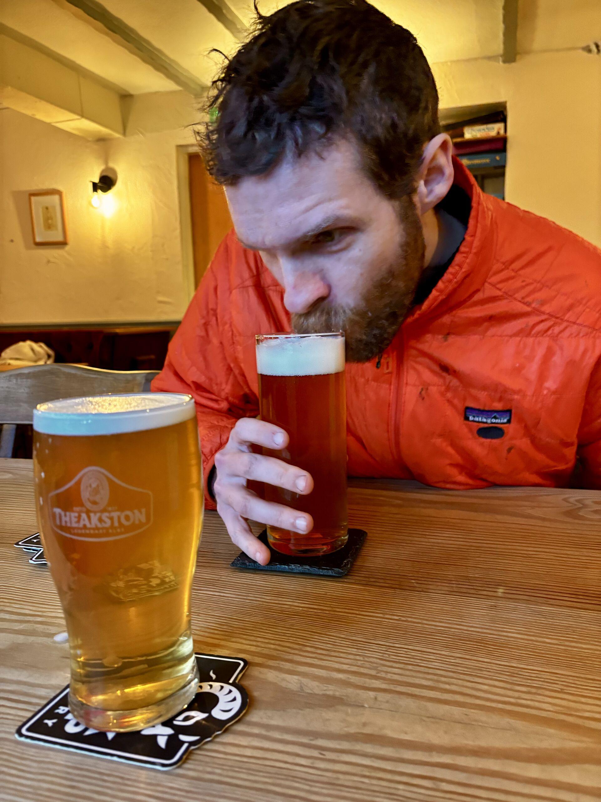 A man in an orange jacket sipping the foam off of a beer; another beer sits on the table in the foreground