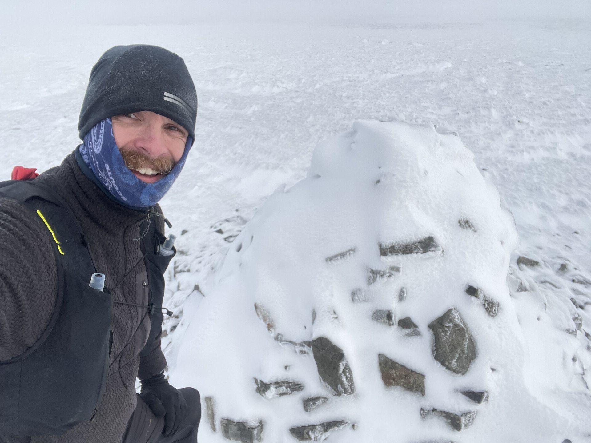 man smiling in a snowy landscape, wearing a beanie and neck gaiter, with a rocky, snow-covered mound next to him.