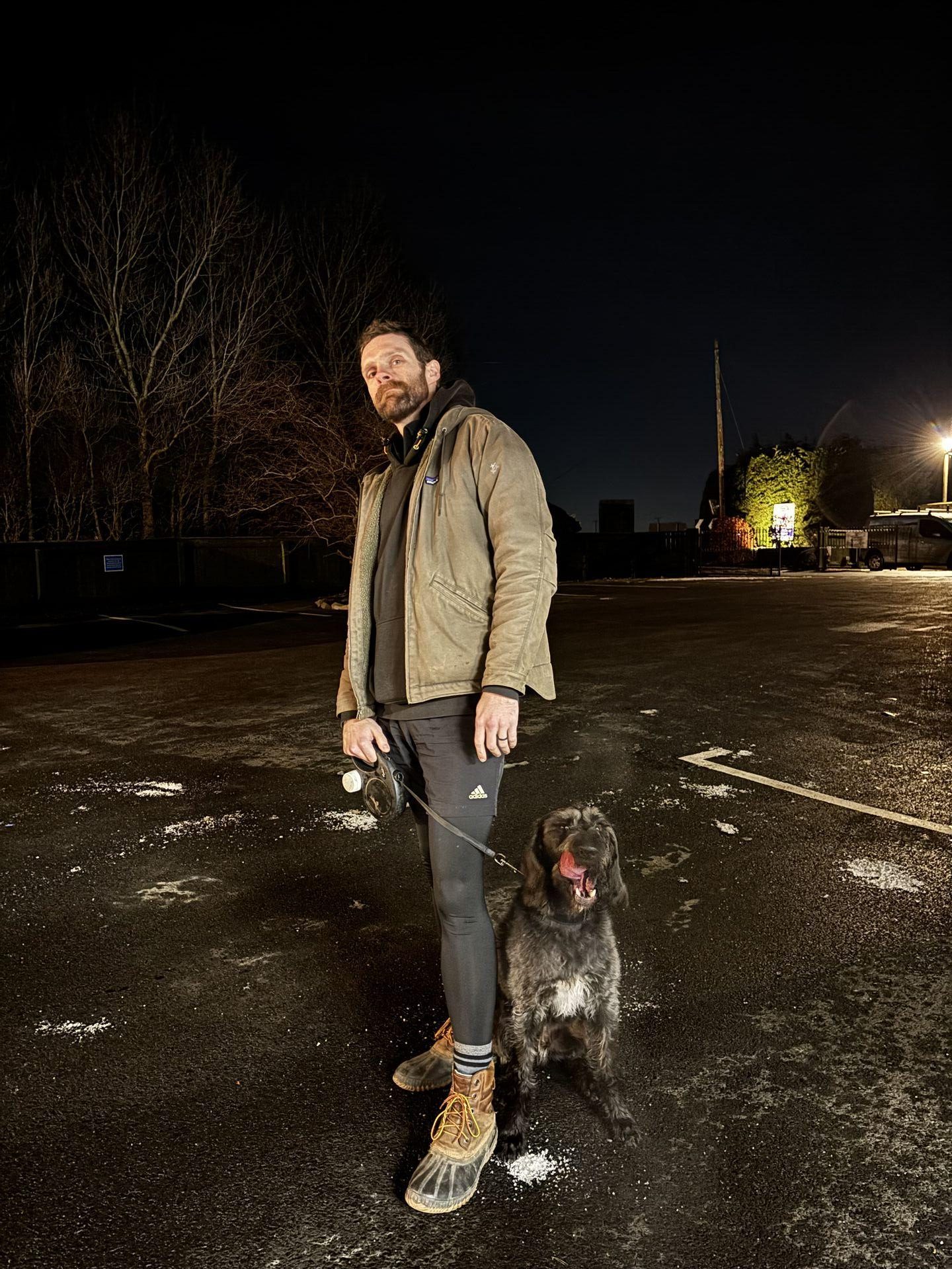 A man in winter jacket, running tights, and LL Bean boots standing in a car park with a scruffy black dog