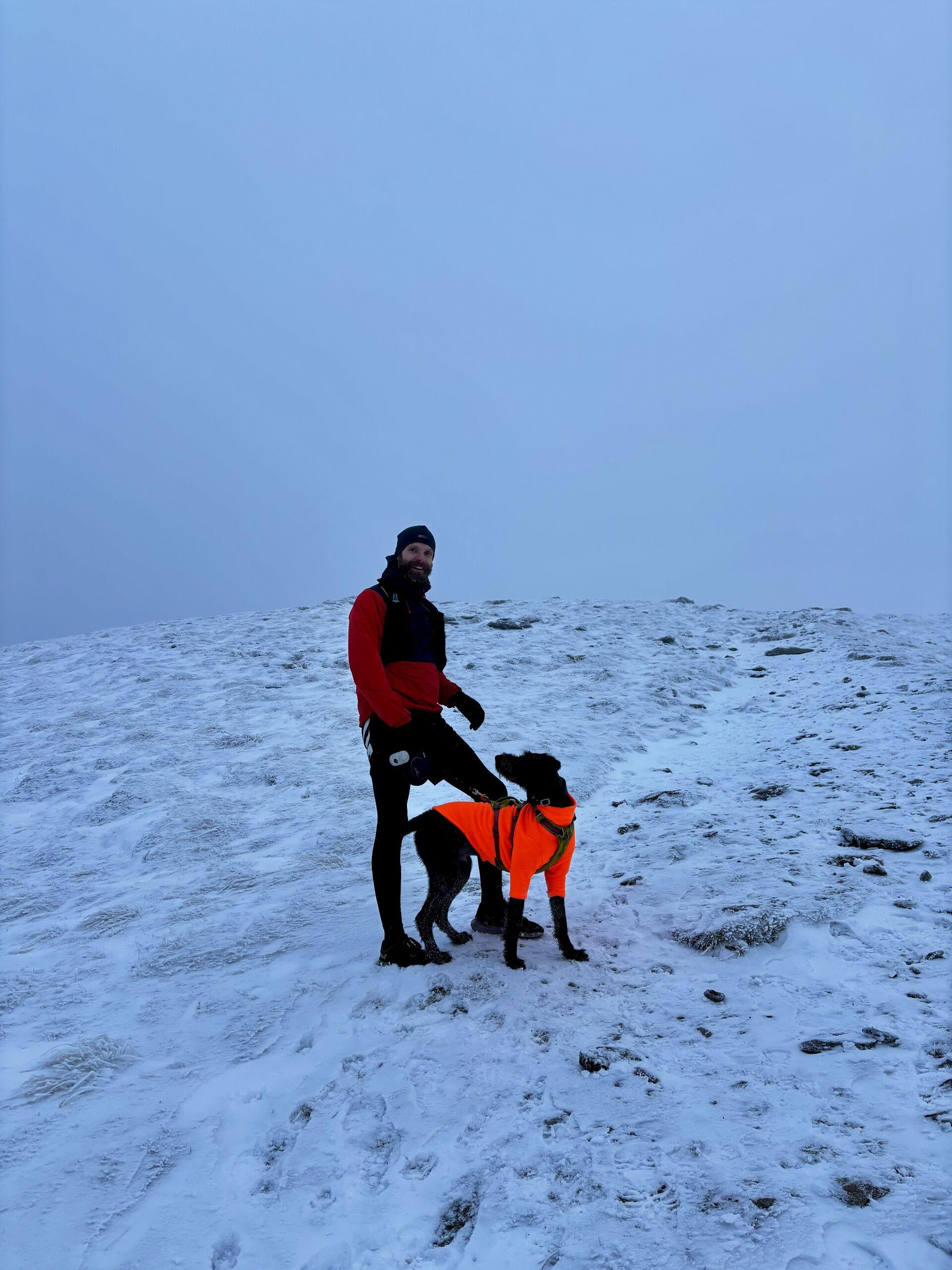 Me in a windbreaker, hat and gloves, standing with Ghyll the dog in an orange fleece, on the side of a bare, snowblown hill