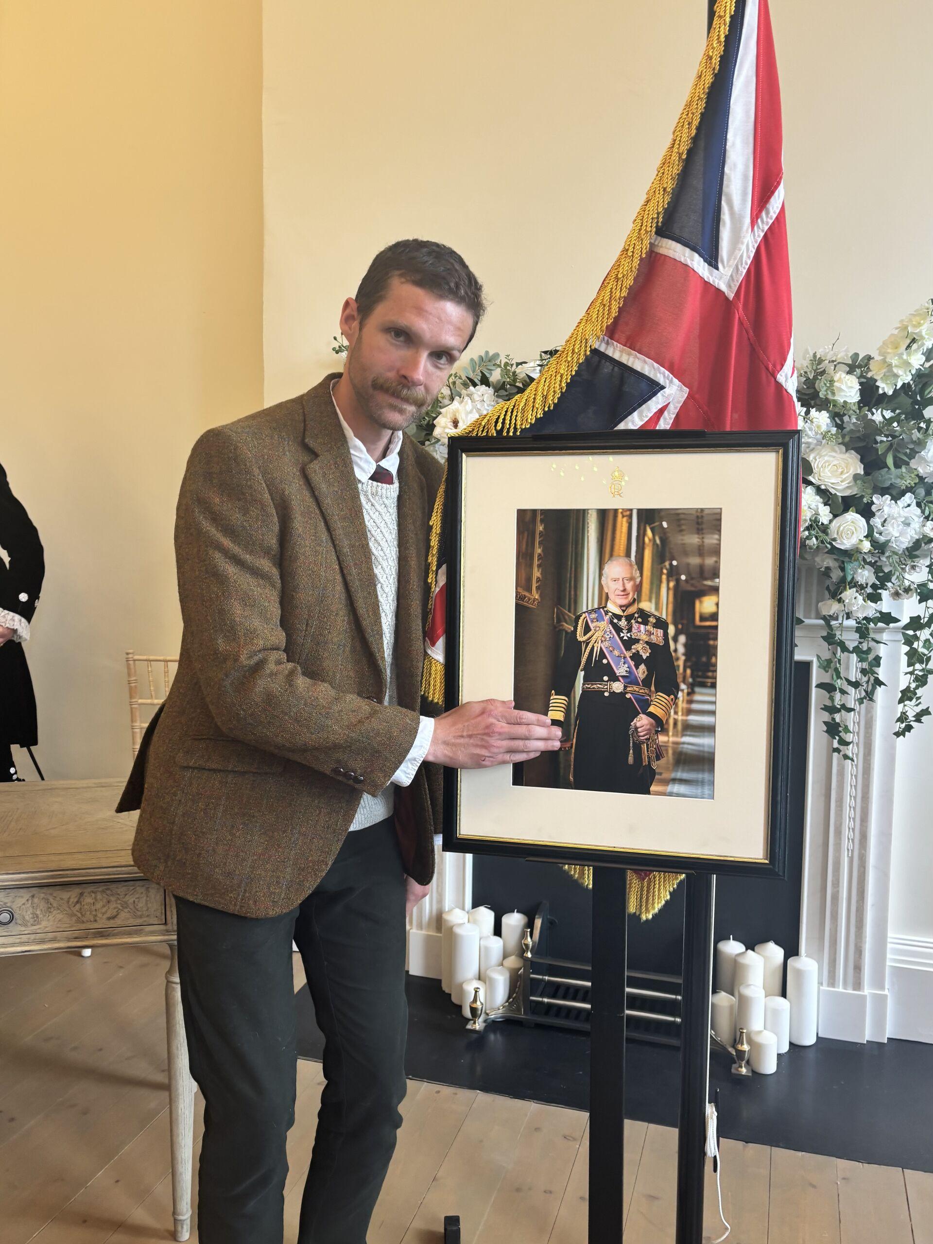 A man in a brown jacket pretending to shake hands with a picture of the king of England, in front of a tasselled Union Jack