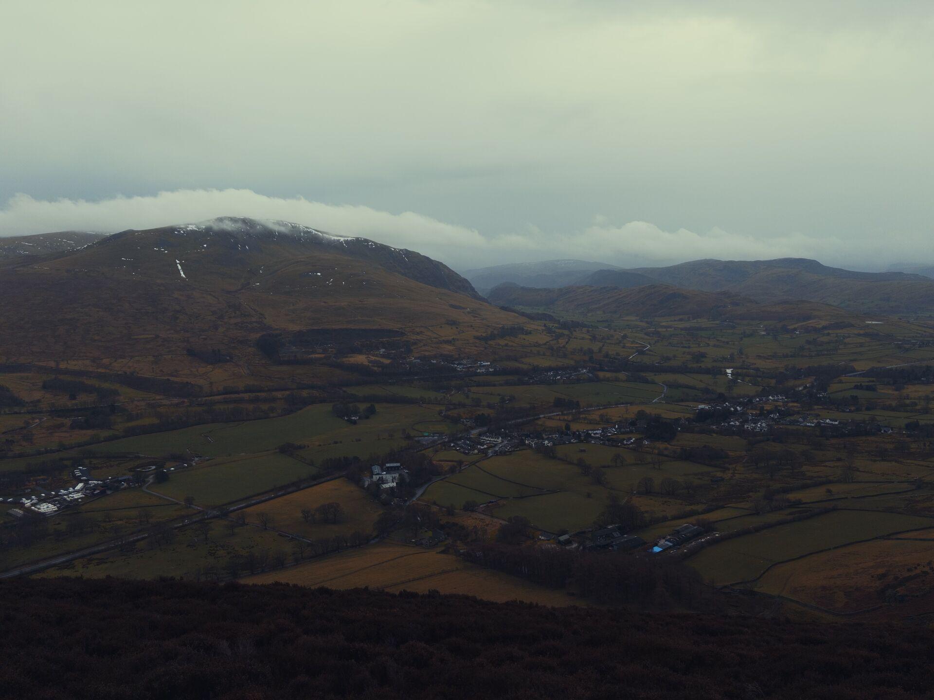 The green fields around Threlkeld and the bulk of Clough Head rising beyond, with some snow remaining on the summit and a halo of cloud
