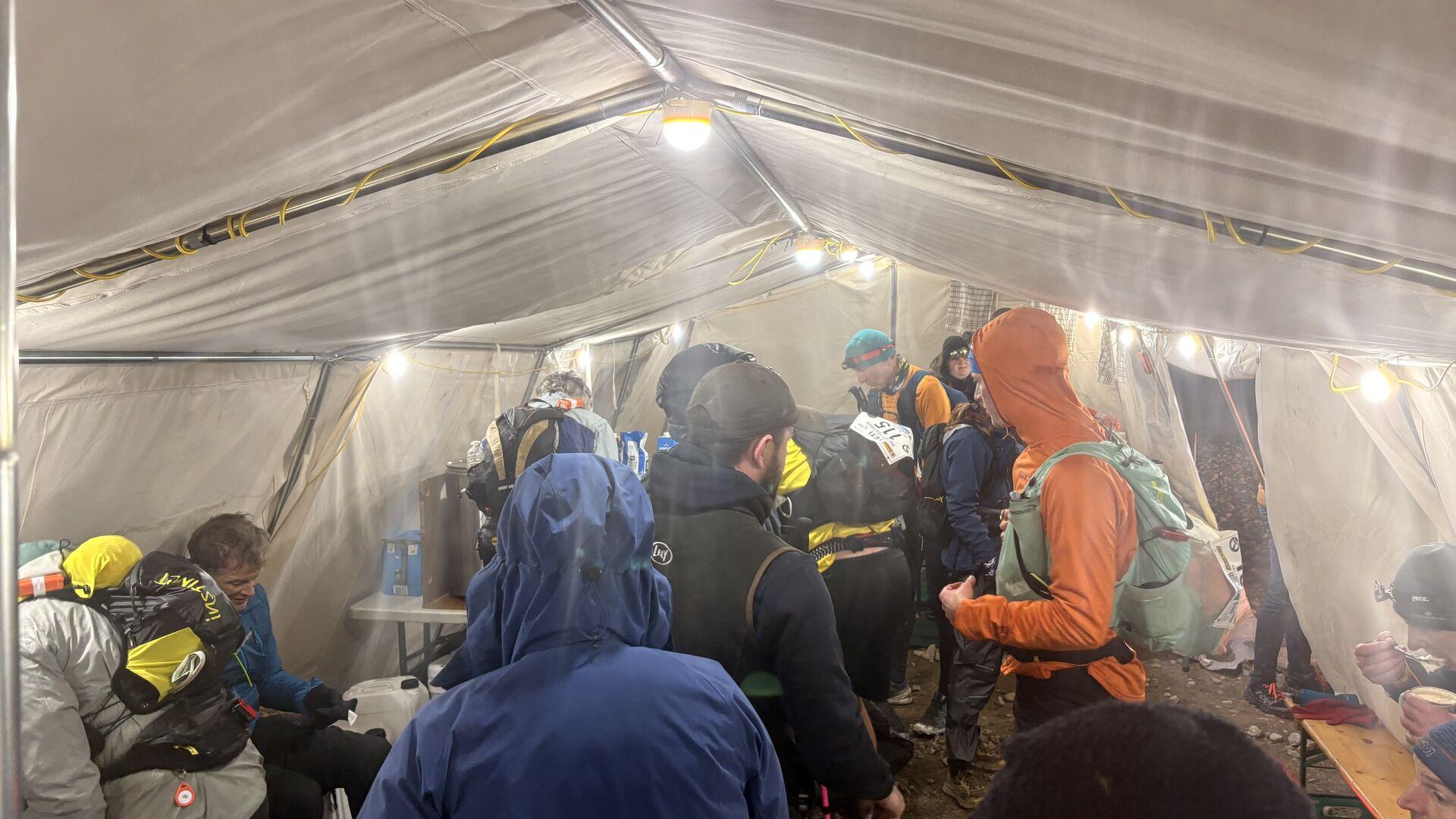 A group of runners in coats alternately sitting and standing inside of a white tent, with bright lights overhead