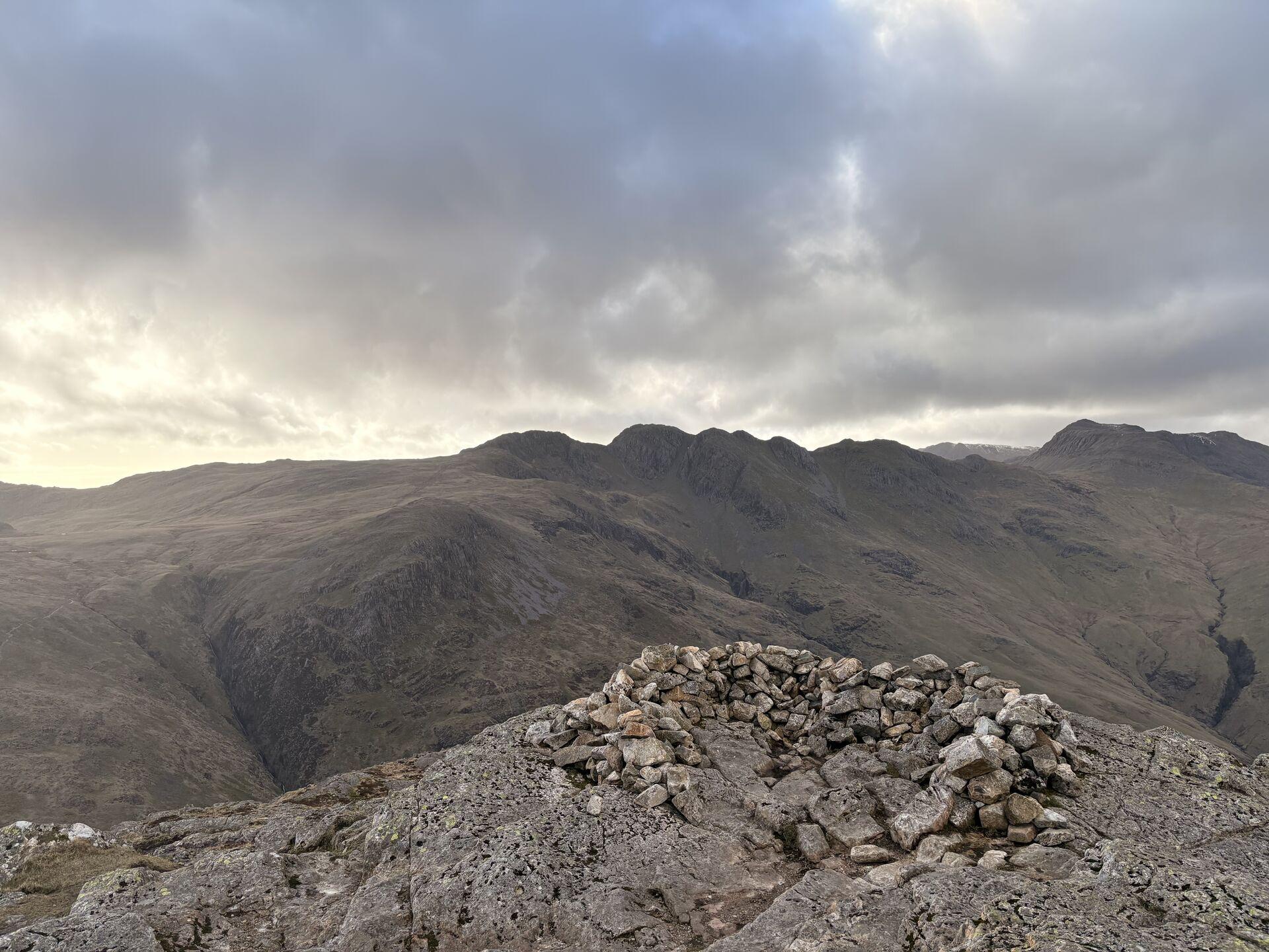 High Lake District fells under a late-day sky; in the foreground is a cairn on top of a rocky summit, beyond an undulating craggy ridgeline, with Bowfell off in the distance on the right