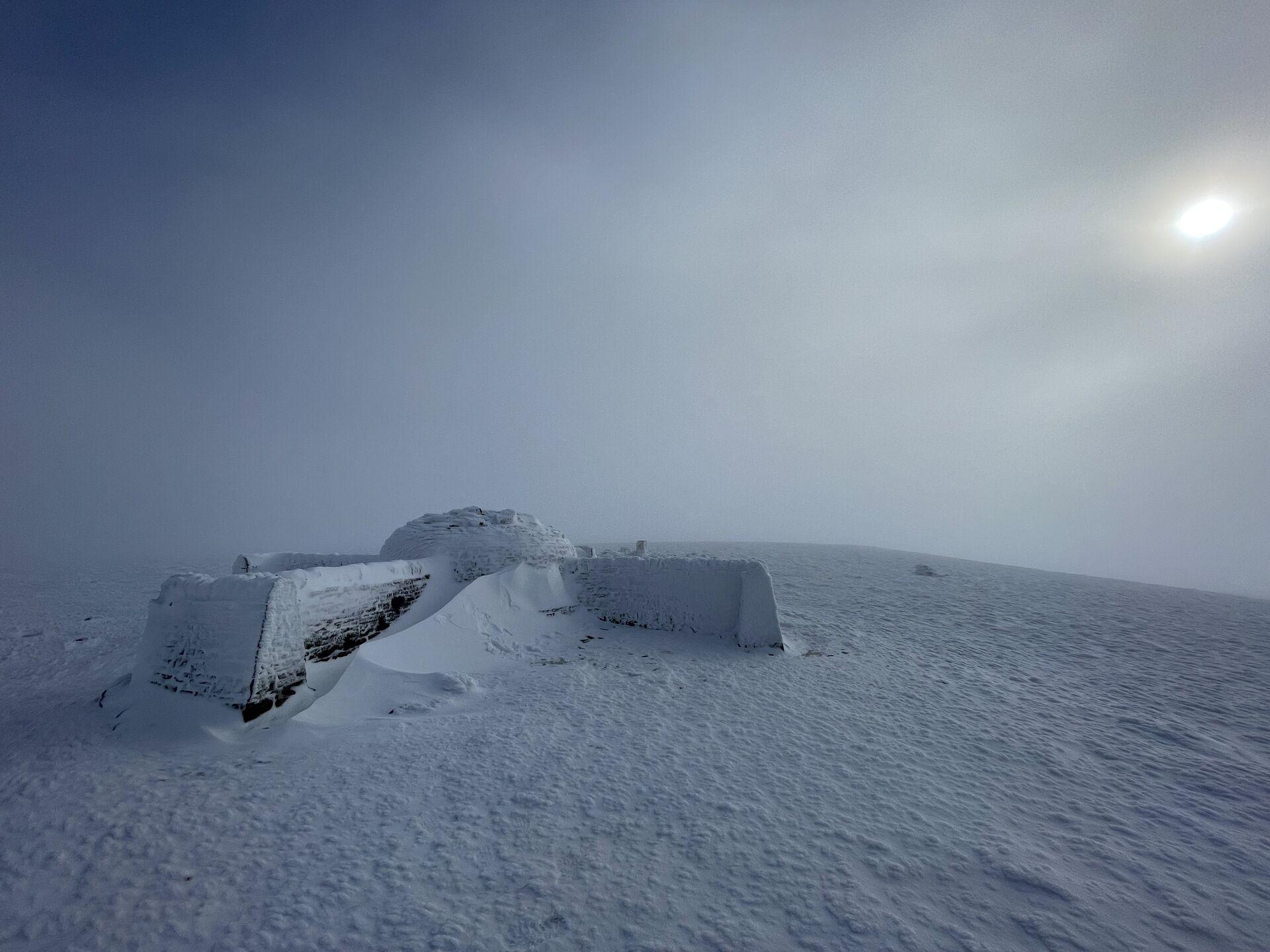 A snowy, windswept landscape under a cloudy sky. In the foreground, there is a structure partially buried in snow, with snowdrifts surrounding it. The scene conveys a sense of isolation and harsh weather conditions, with a faint sun visible in the background.