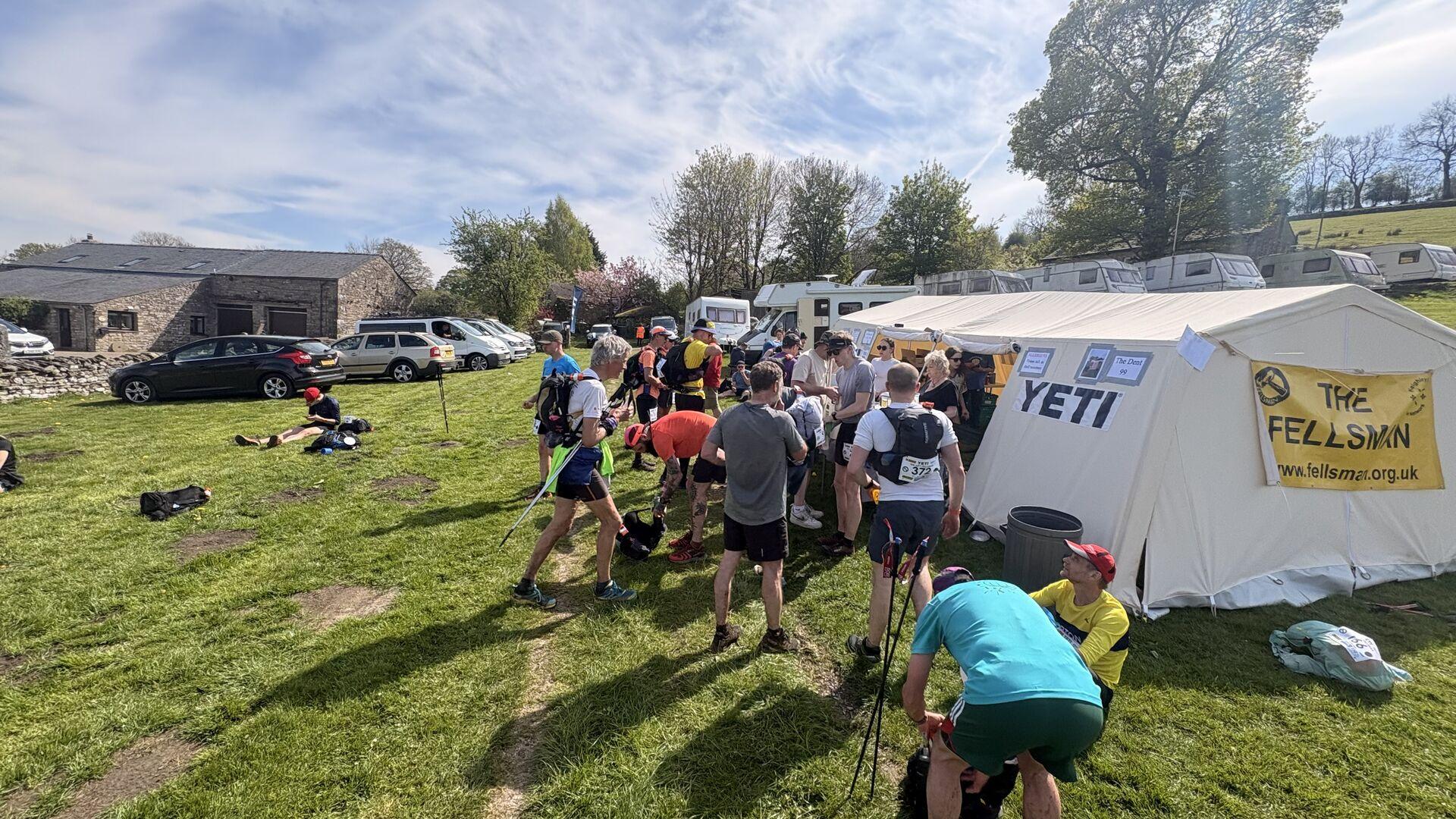 A group of runners standing idly around a white marquee in a grassy field, with some cars parked off to the side and trees behind