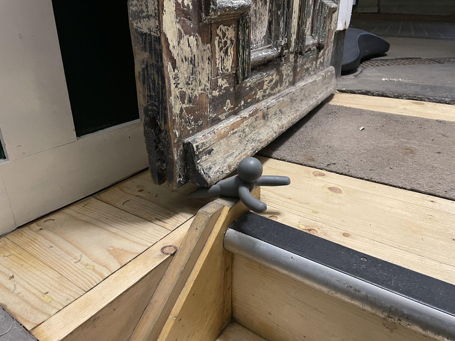 A gray doorstop shaped like a small figure is wedged against a rustic wooden door, partially open. The door is weathered with peeling paint, and the floor features wooden planks and a carpeted area.