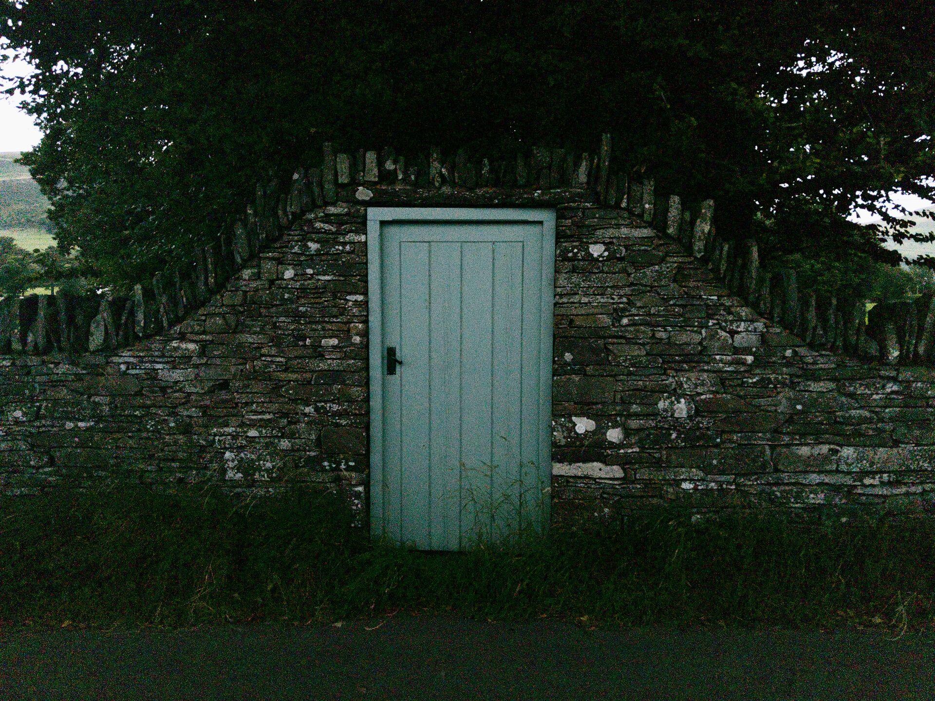 A pale blue wooden door set into a dry stone wall, with trees visible above the wall and open countryside behind.