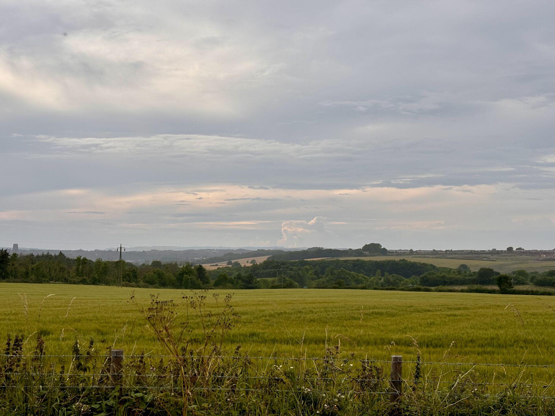 A cloudy sunset over County Durham, with low rolling hills in the foreground, wooded valleys in the distance, and tall thunderclouds off near the horizon in the orange dusk light