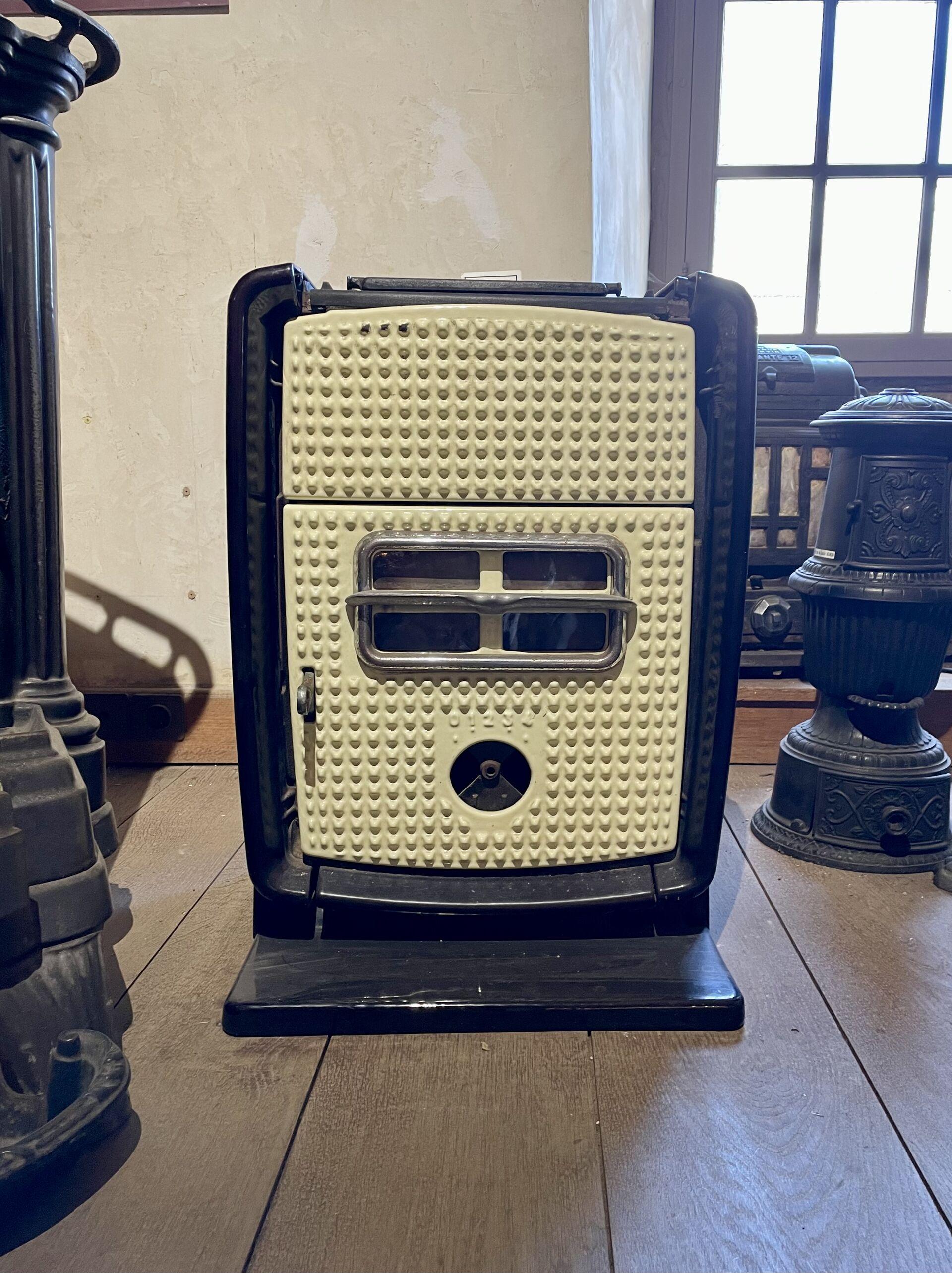 A cream-fronted enamelware stove with chrome accents and black sides, on a wooden floor, surrounded by small cast iron stoves