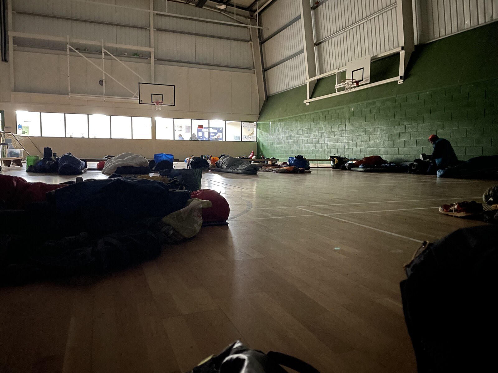 A bunch of people all curled up in sleeping bags on the floor of a gymnasium. The lights are out in the gym but the light from the hall floods through the glass at the end of the room.