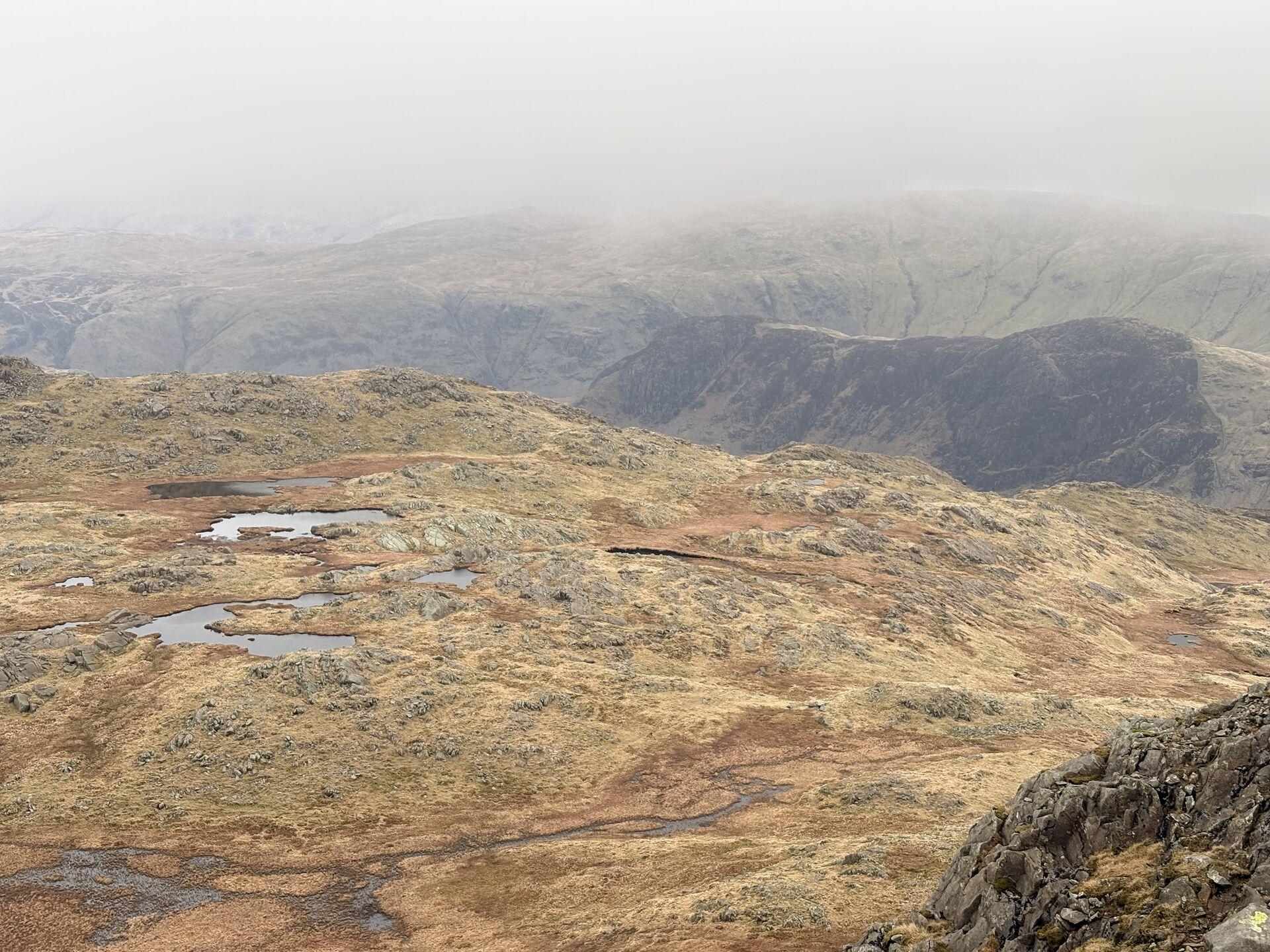 A few small tarns in a brown-grey plateau of low grass and crag, with some Lakeland fells extending into the distance beyond, fading into low cloud.