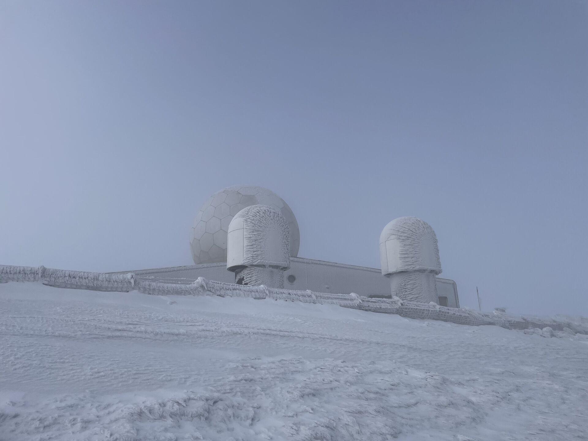 A radar station located on the summit of a mountain, surrounded by a snowy landscape. The building features two large spherical structures covered in frost. The sky is overcast and grey, indicating cold weather, while snow blankets the ground. The scene conveys a remote and wintry atmosphere.