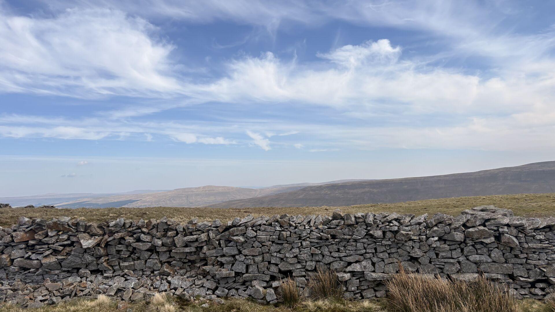 A view of a wispy clouded sky over rolling brownish hills, with a stone wall across the bottom of the frame