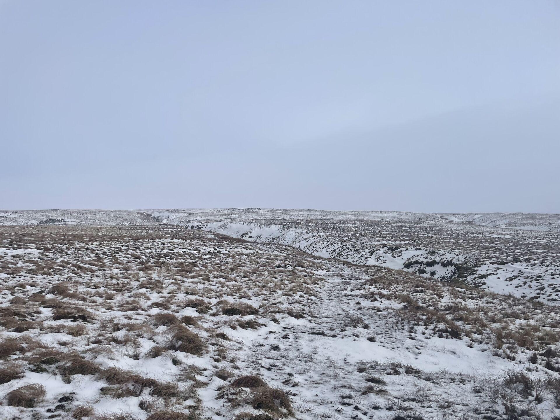 Snow-covered landscape with sparse grass and a gentle slope cut across by a straight ditch, under a grey sky.