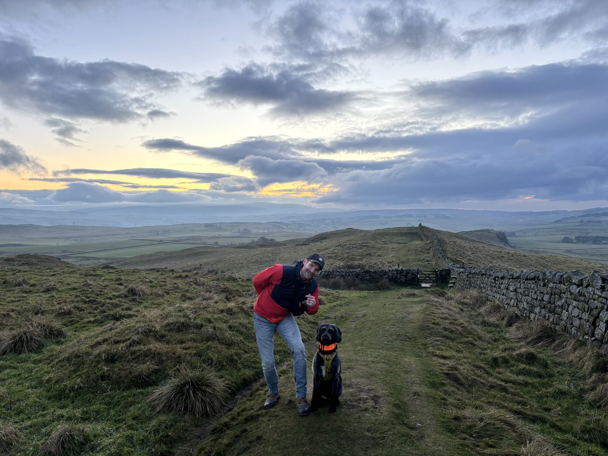 A man in a red jacket and a black scruffy dog sitting next to him, posing on a grassy hilltop with a stone wall running off over the curve of the hill on the right, with a darkening sky behind