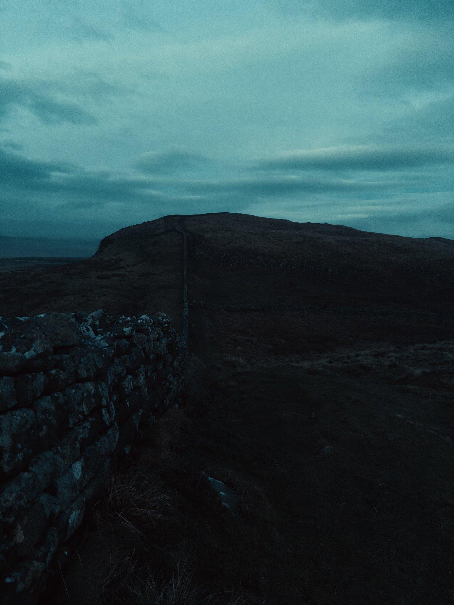A stone wall runs down a craggy hillside and back up the hill on the far side, with a dark cloudy sky above