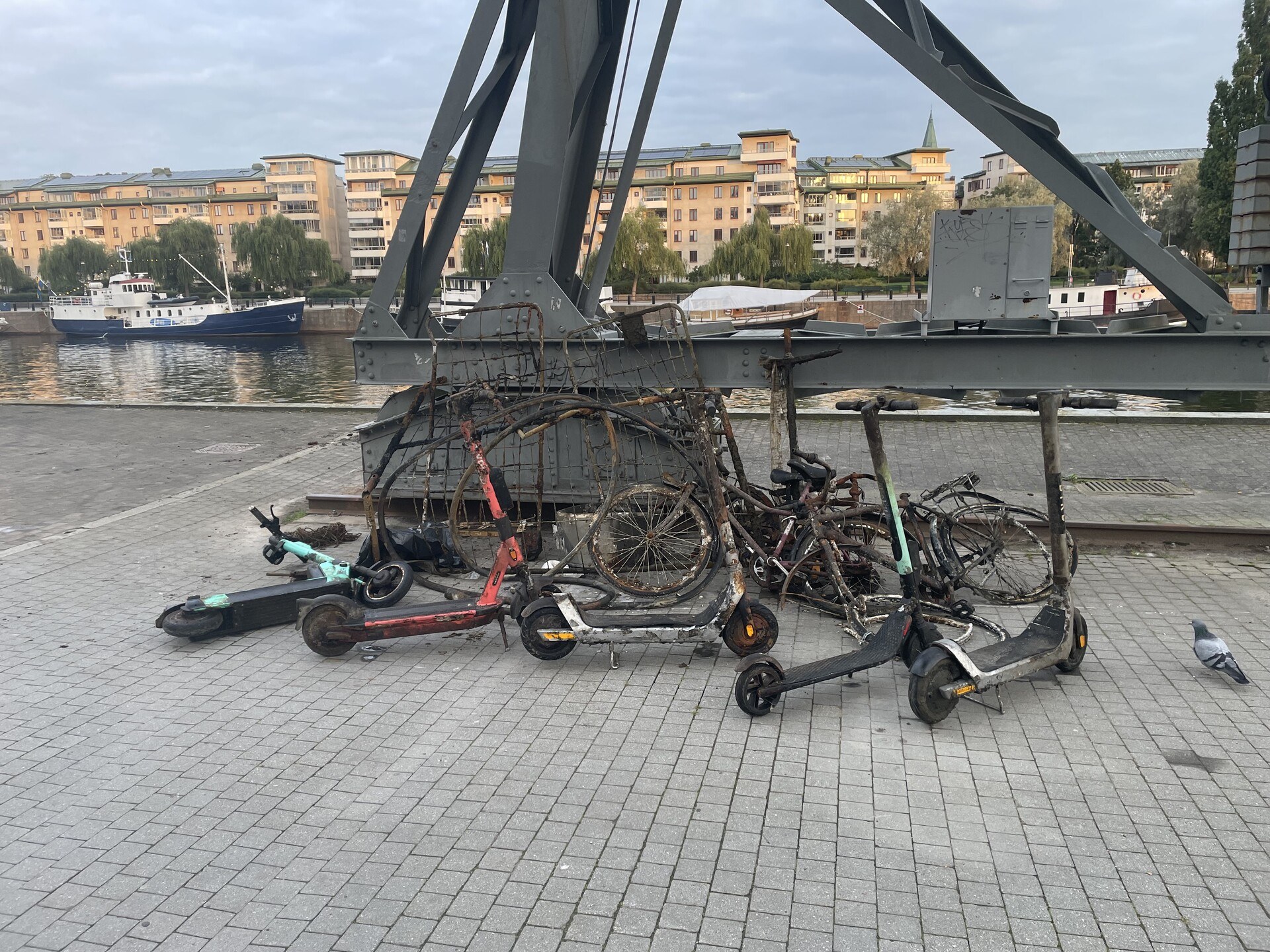 A handful of rusty bikes and grimy electric scooters left by the iron legs of an old fixed crane on the canalside