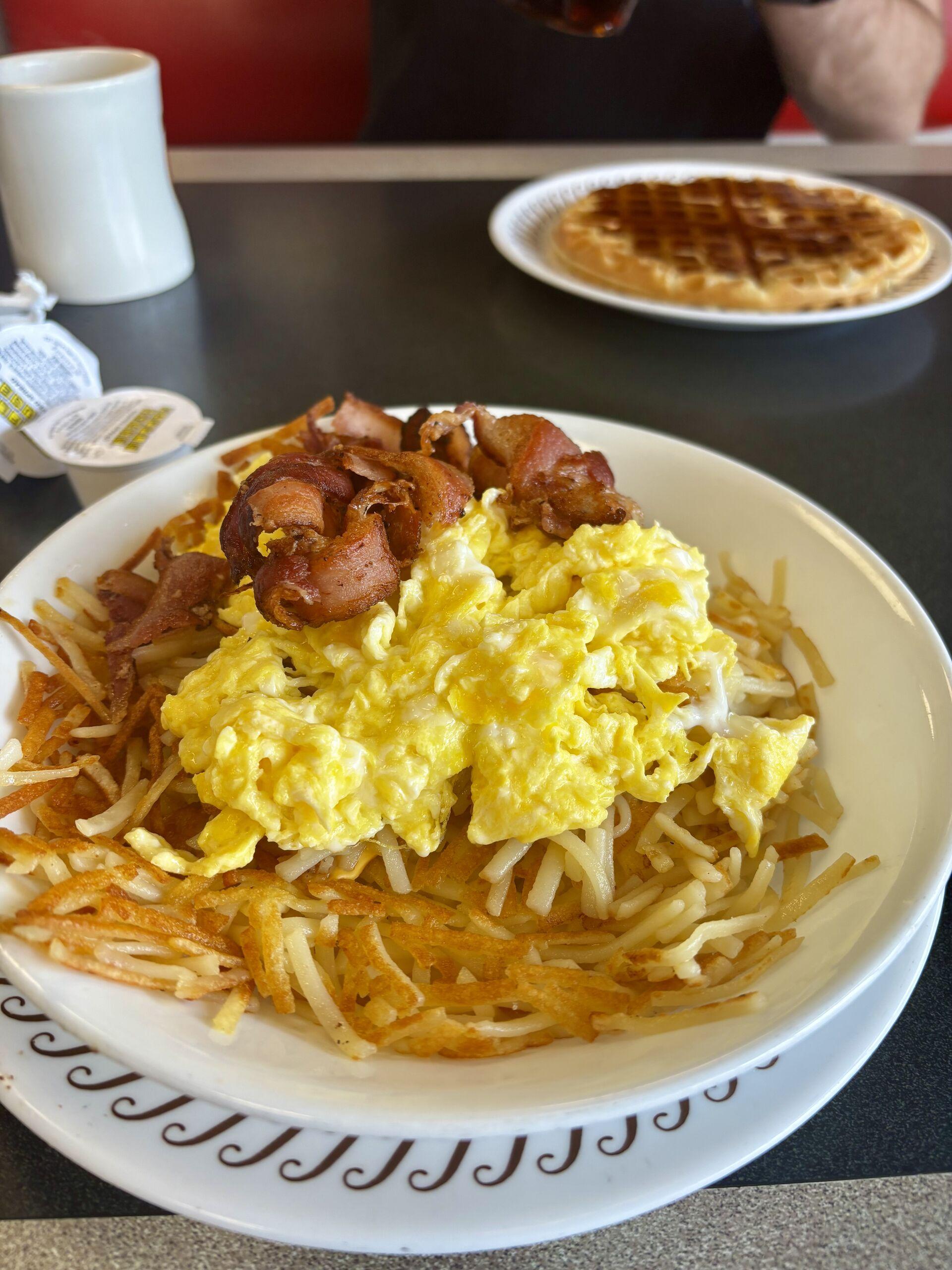 A bowl of hashbrowns topped with American cheese, scrambled eggs, and small slices of bacon