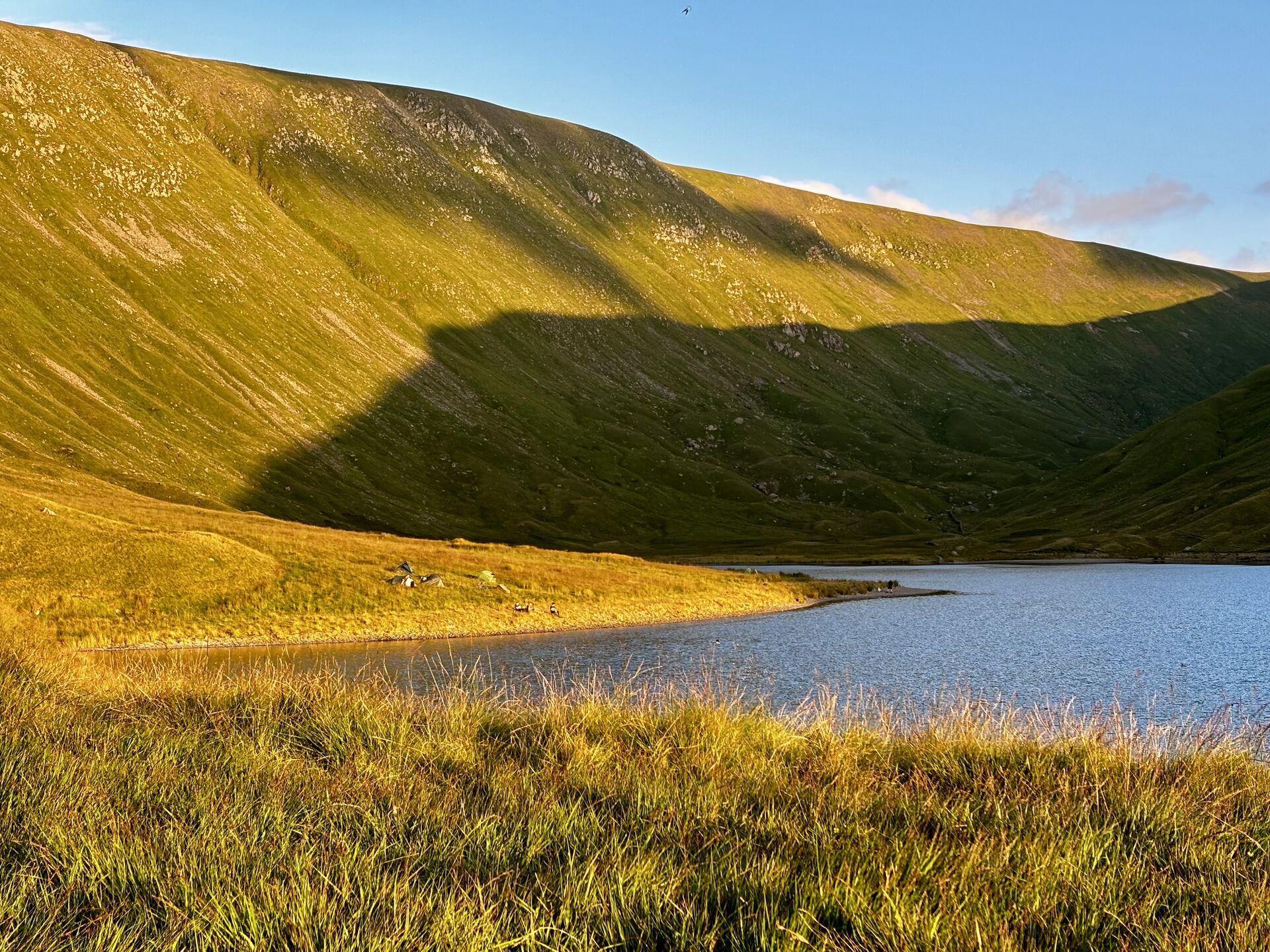 Grassy hillside leading down to a lake, with sunset shining on half the hillside and the other half in the shadow of the opposing hill, with some short grasses in the immediate foreground