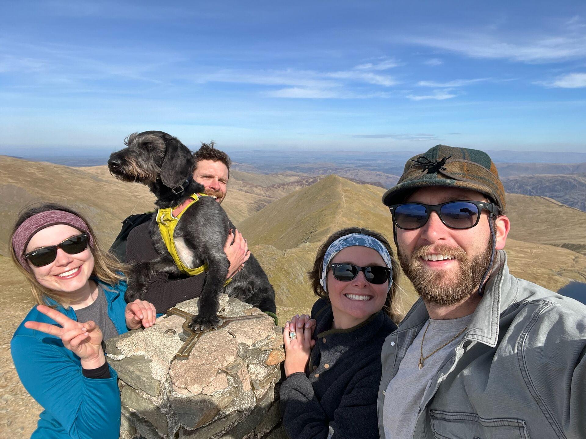 Four people and a dog posing for a selfie at the summit of Helvellyn, with mountains stretching off in the background